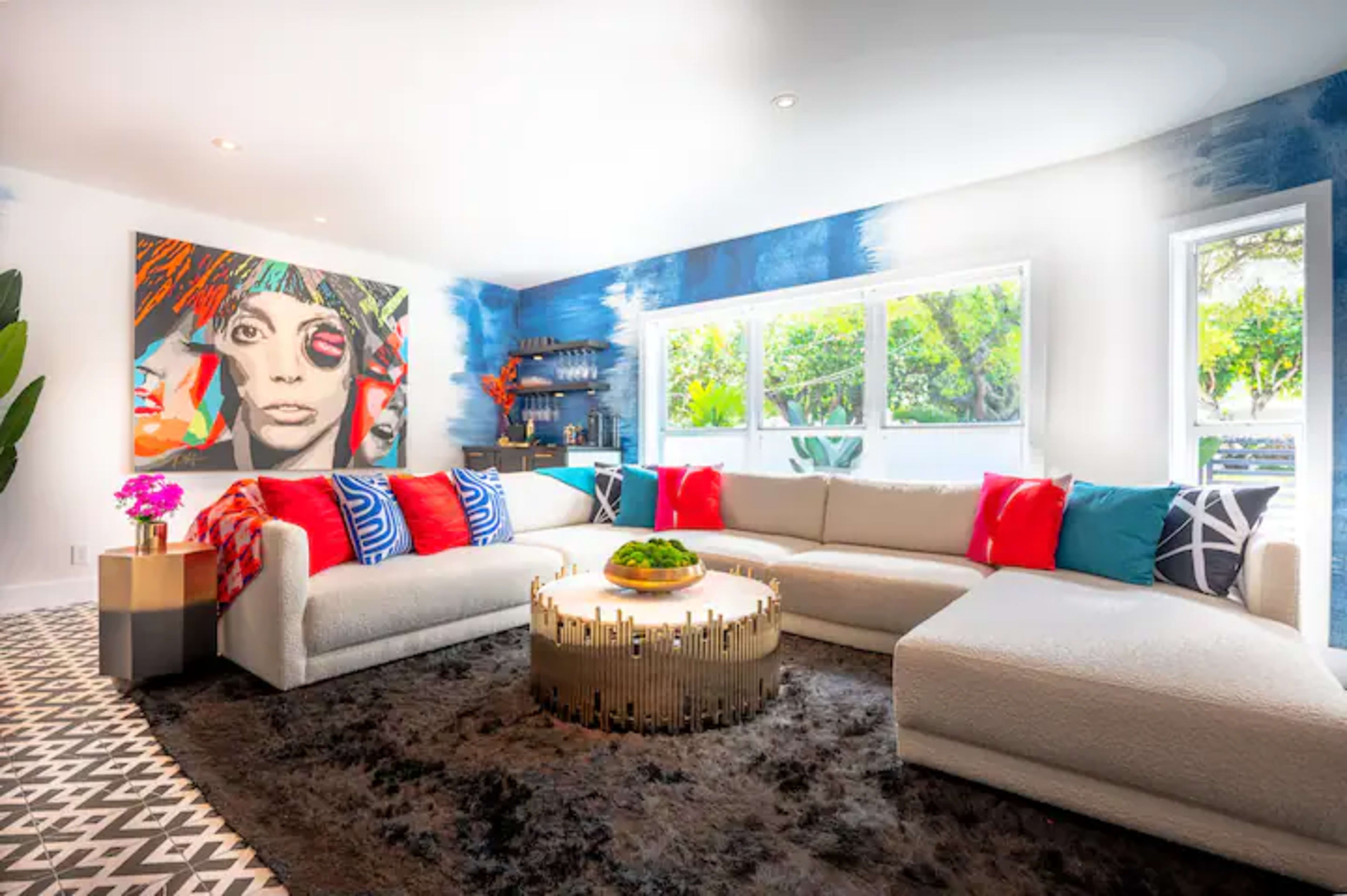 A modern living room features a large sectional sofa with colorful cushions, a circular coffee table, and a vibrant wall painting.