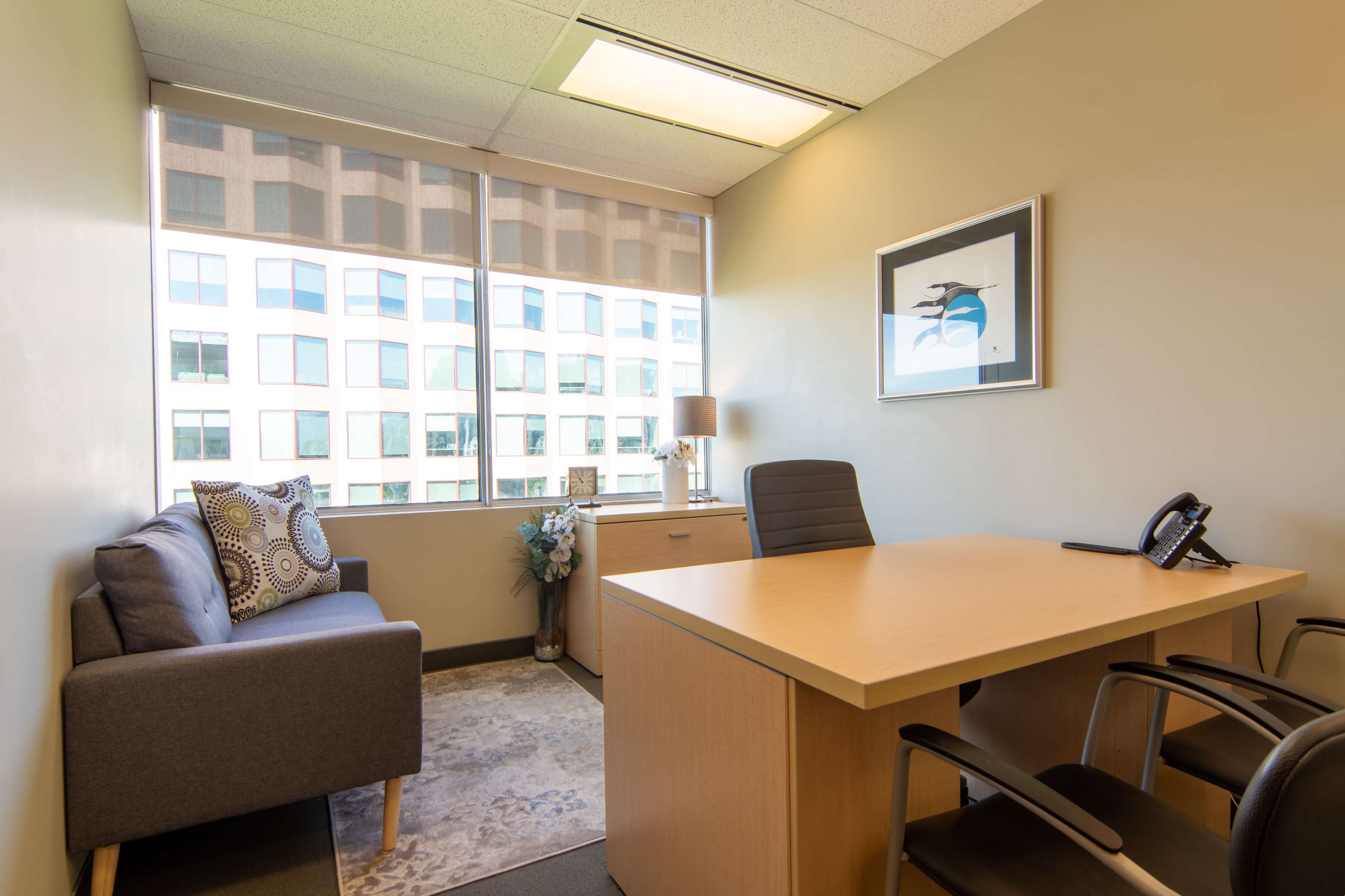 The image shows a well-lit office space featuring a desk, a chair, a couch, and a large window with a view of an exterior building.