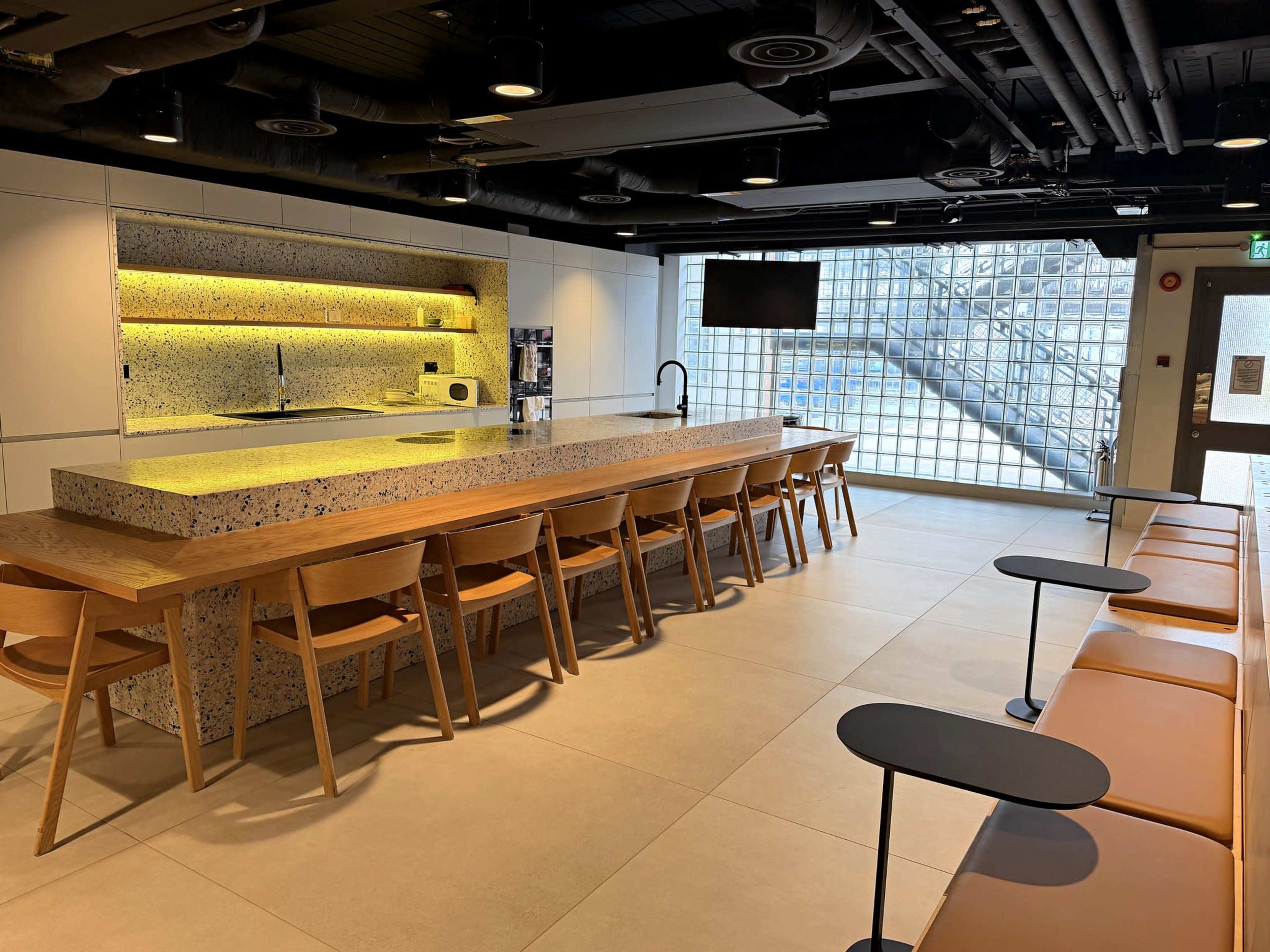 The image shows a modern cafe-style interior with a long communal table, wooden chairs, a stone countertop, and a large window with glass blocks.