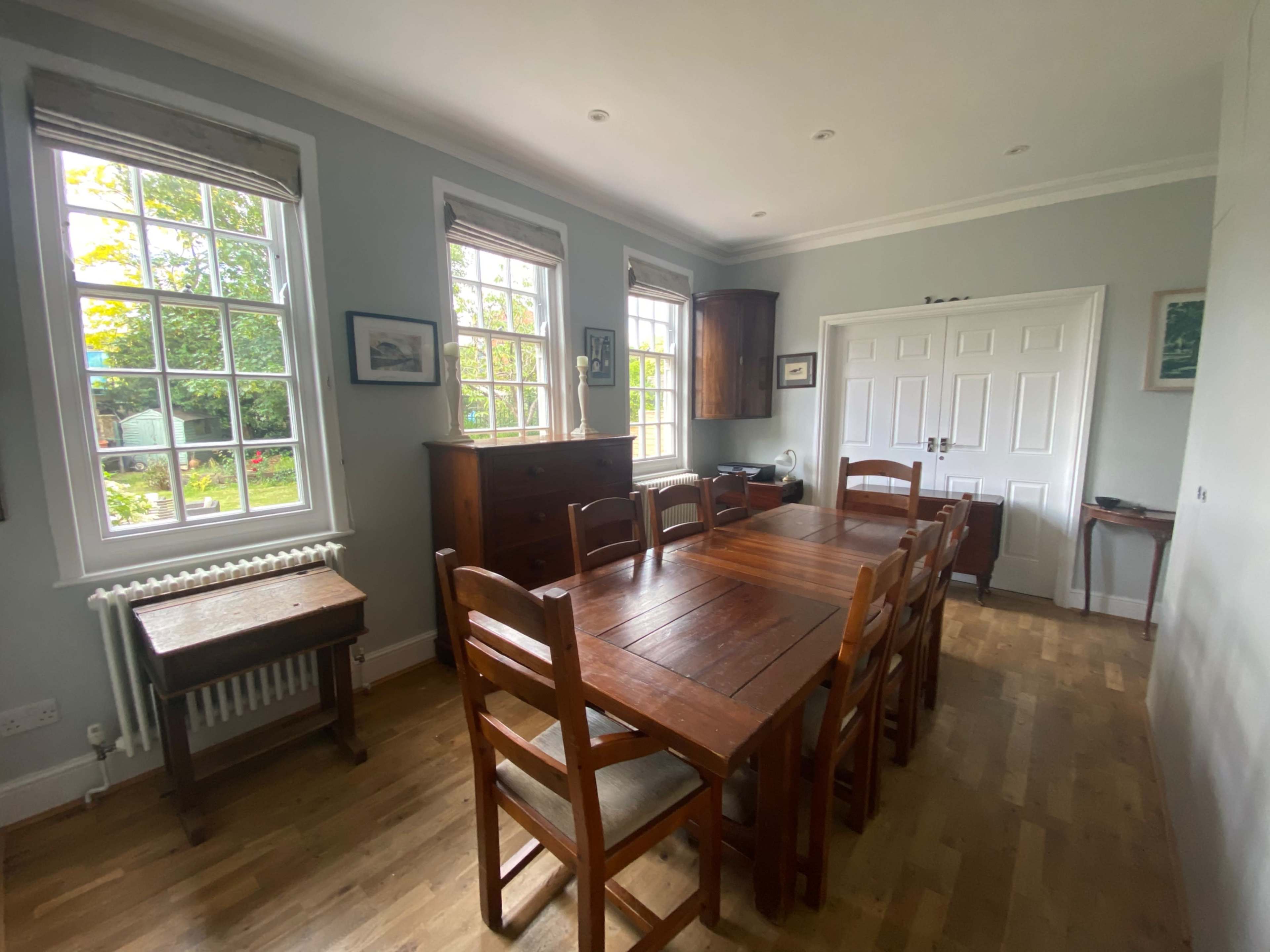 A dining room with a wooden table and chairs, large windows, and a sideboard against the wall.