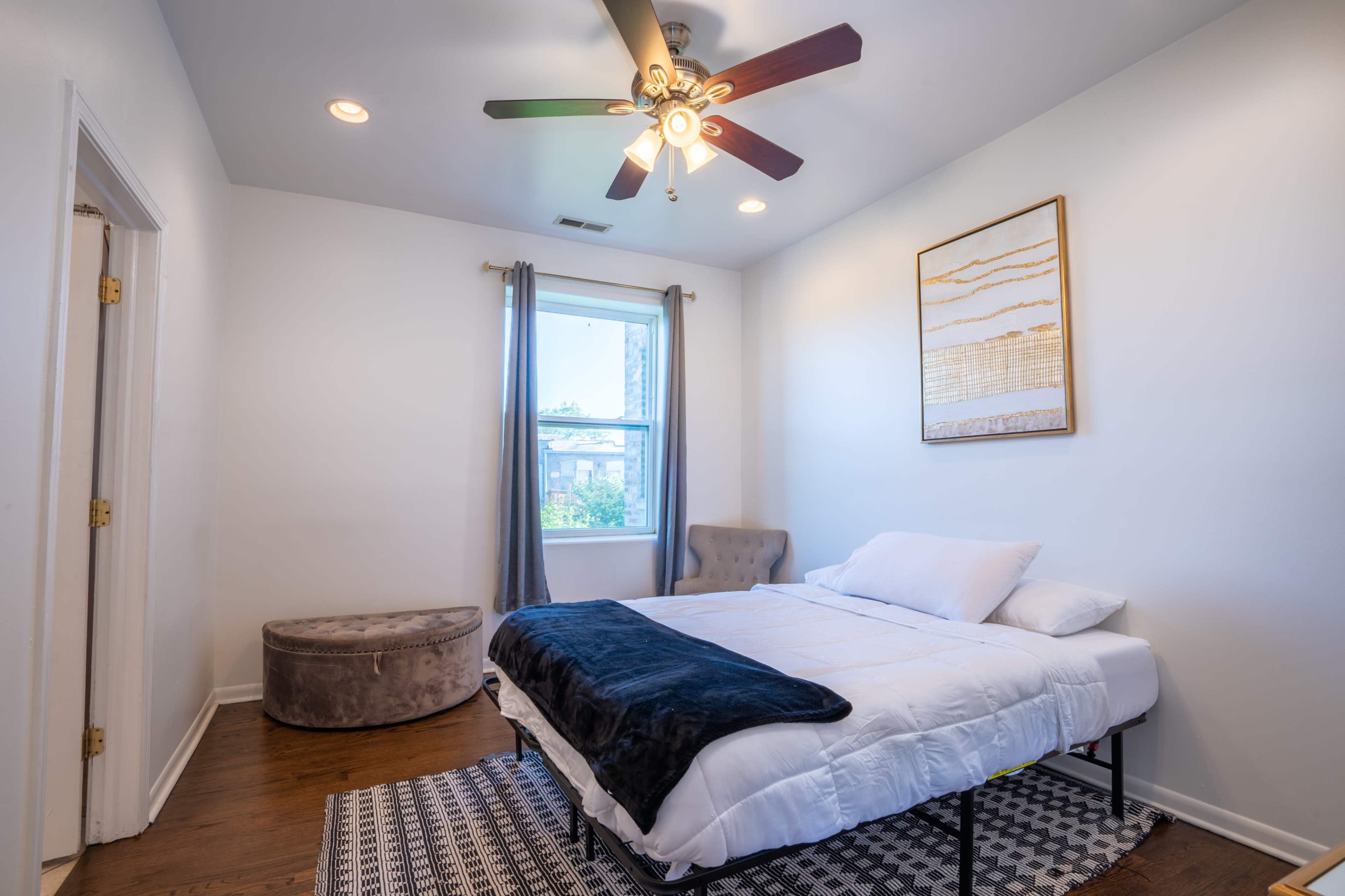 A bedroom with a bed made up in the center, a window with curtains, a decorative wall hanging, a chair in the corner, and a round ottoman.