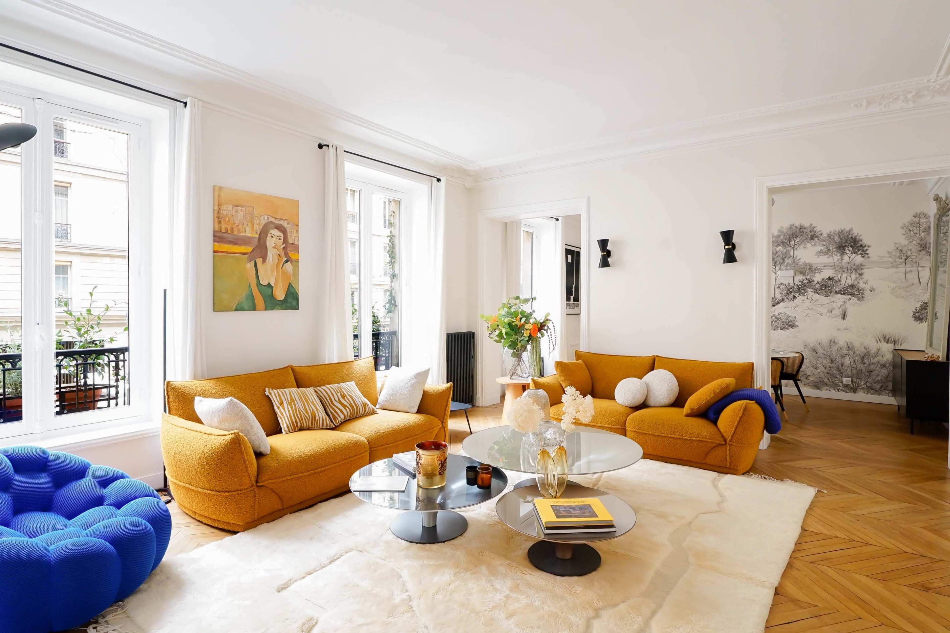 A bright living room features two yellow sofas, a blue circular chair, a glass coffee table, and large windows that allow natural light to enter.