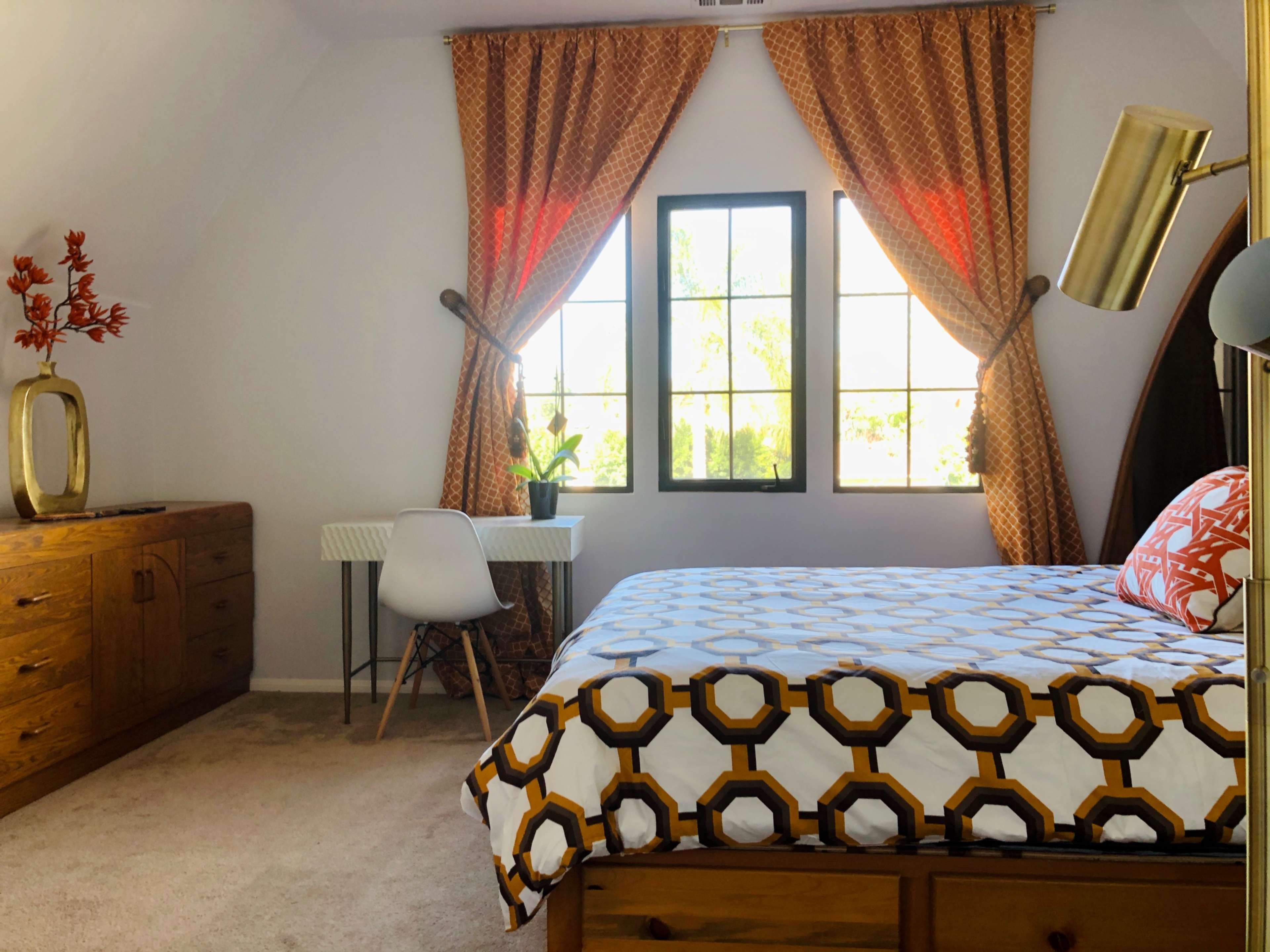 A bedroom featuring a bed with a patterned white and orange blanket, a wooden dresser, a small desk by a window, and curtains in an orange hue.