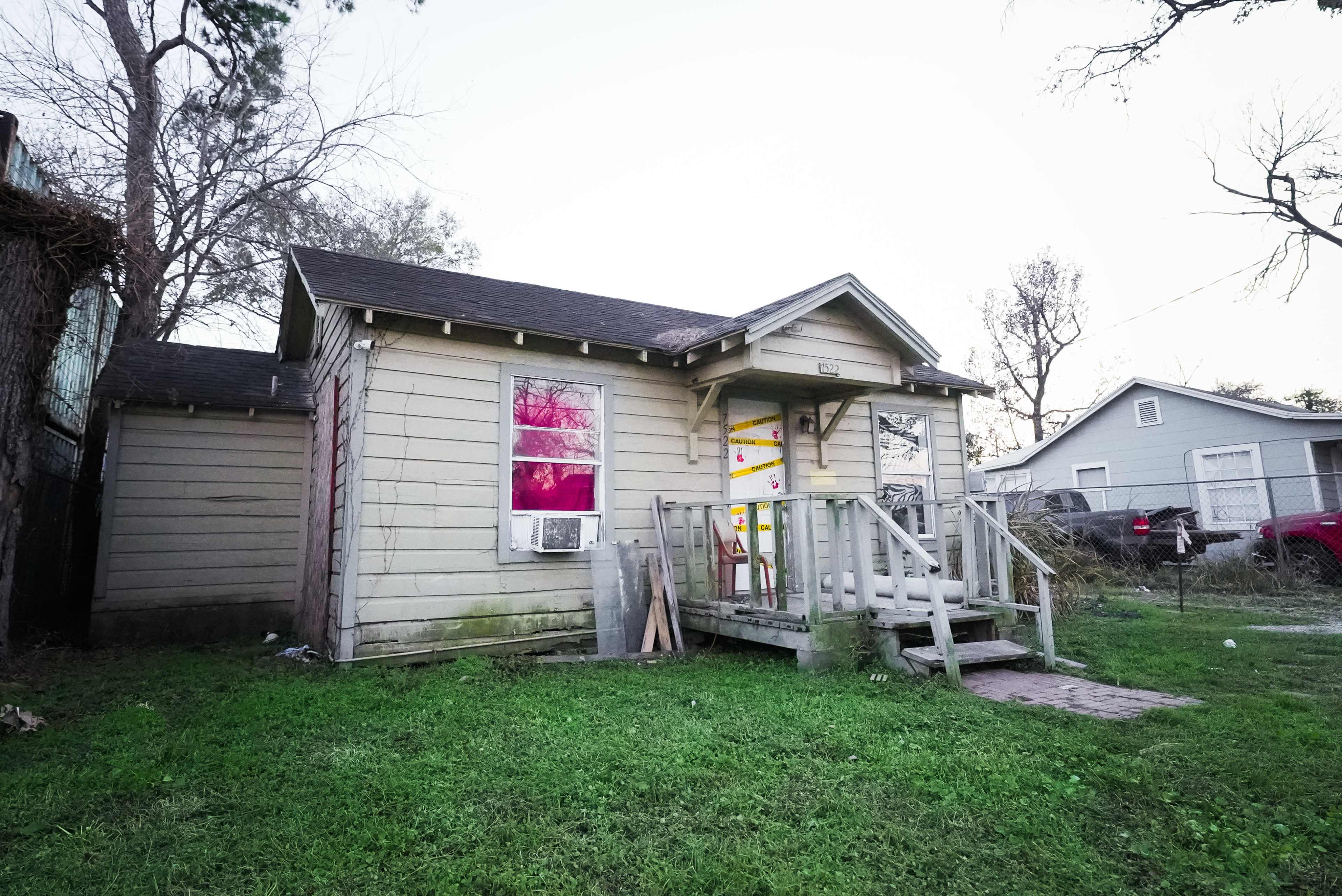 The image shows a small, single-story wooden house with a porch and a few visible boarded windows, surrounded by grass and trees.