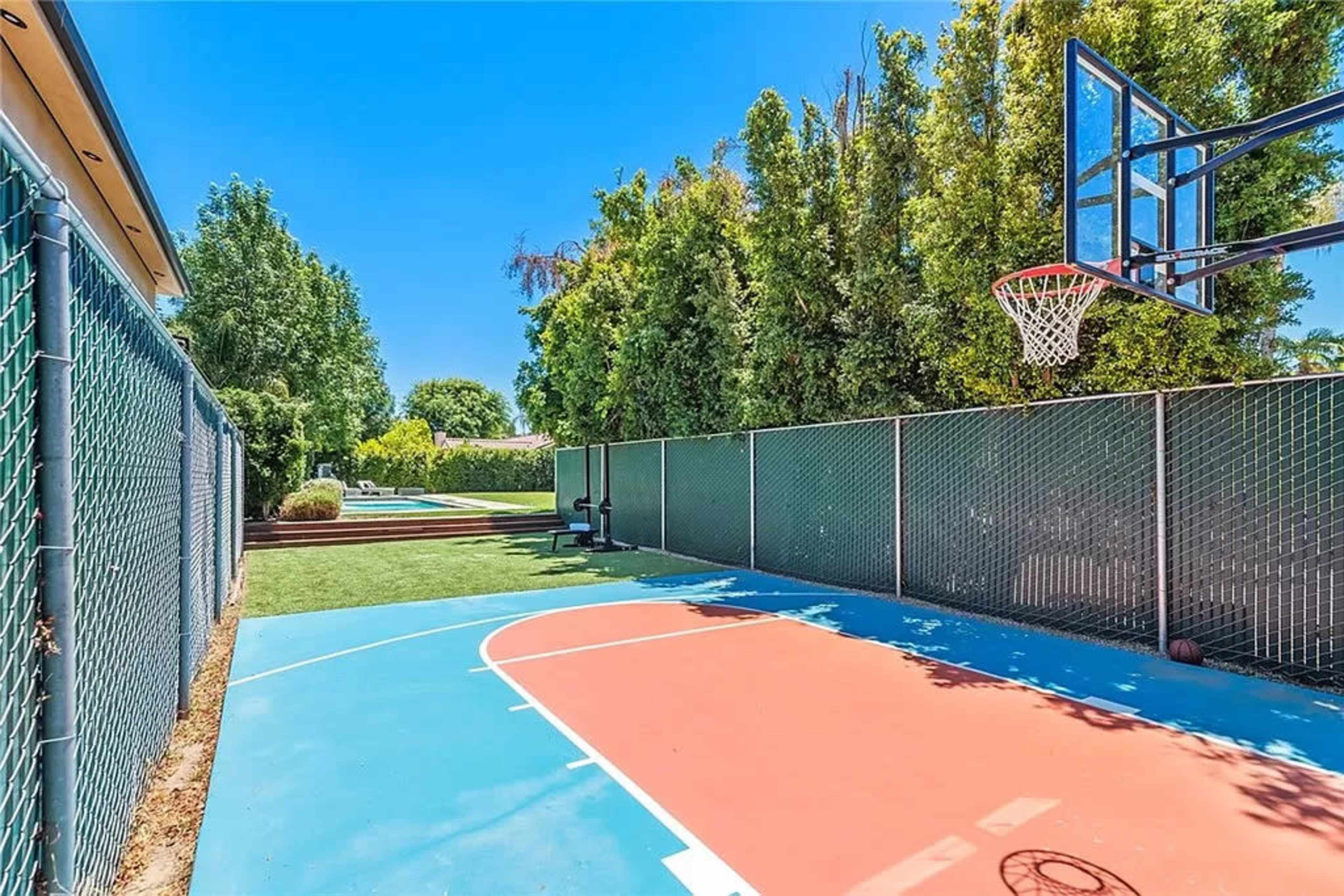 A outdoor basketball court featuring orange and blue sections, surrounded by chain link fencing and lush greenery.