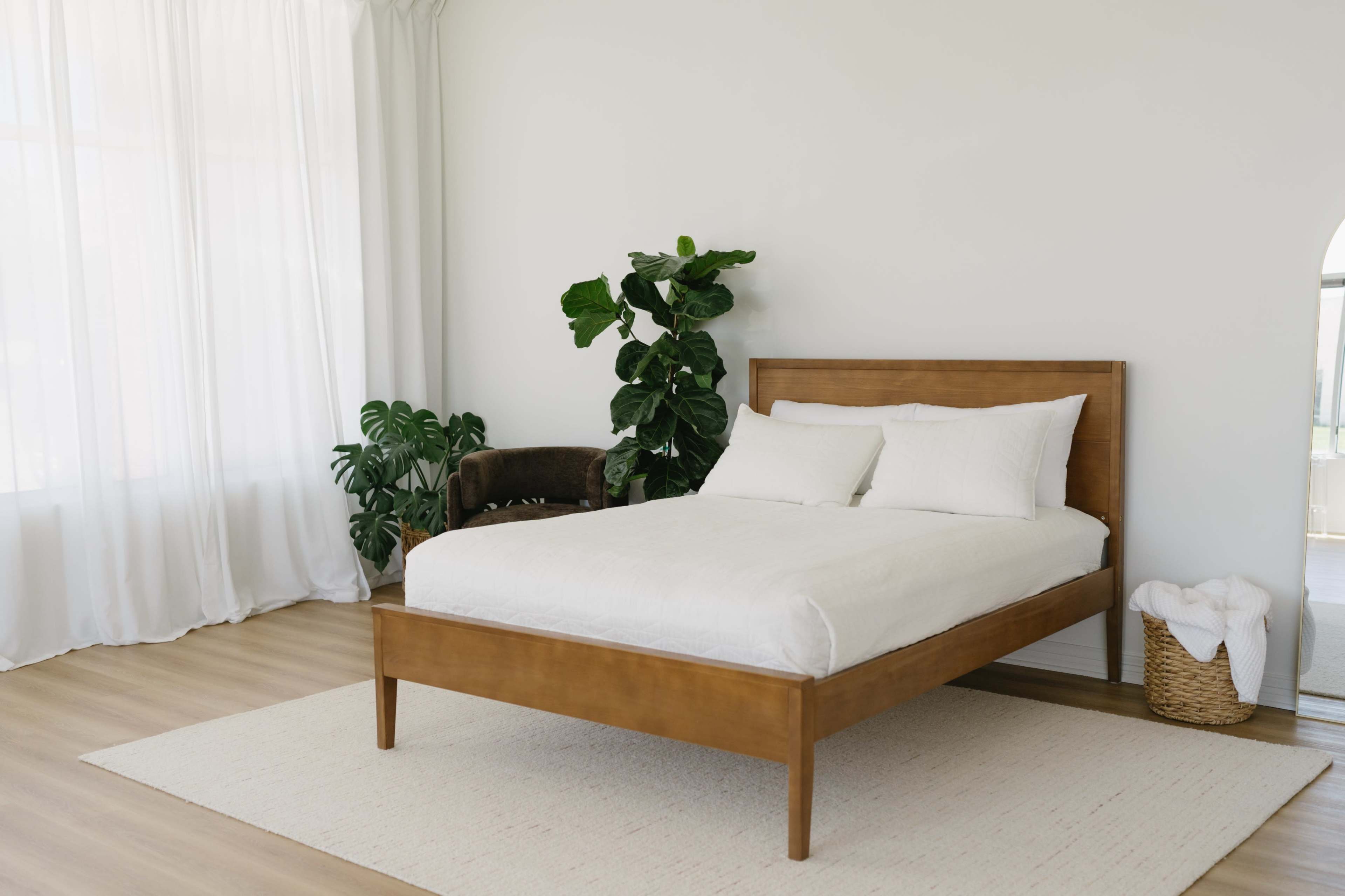 The image shows a minimalist bedroom with a wooden bed, white bedding, a plant, and light-colored curtains.