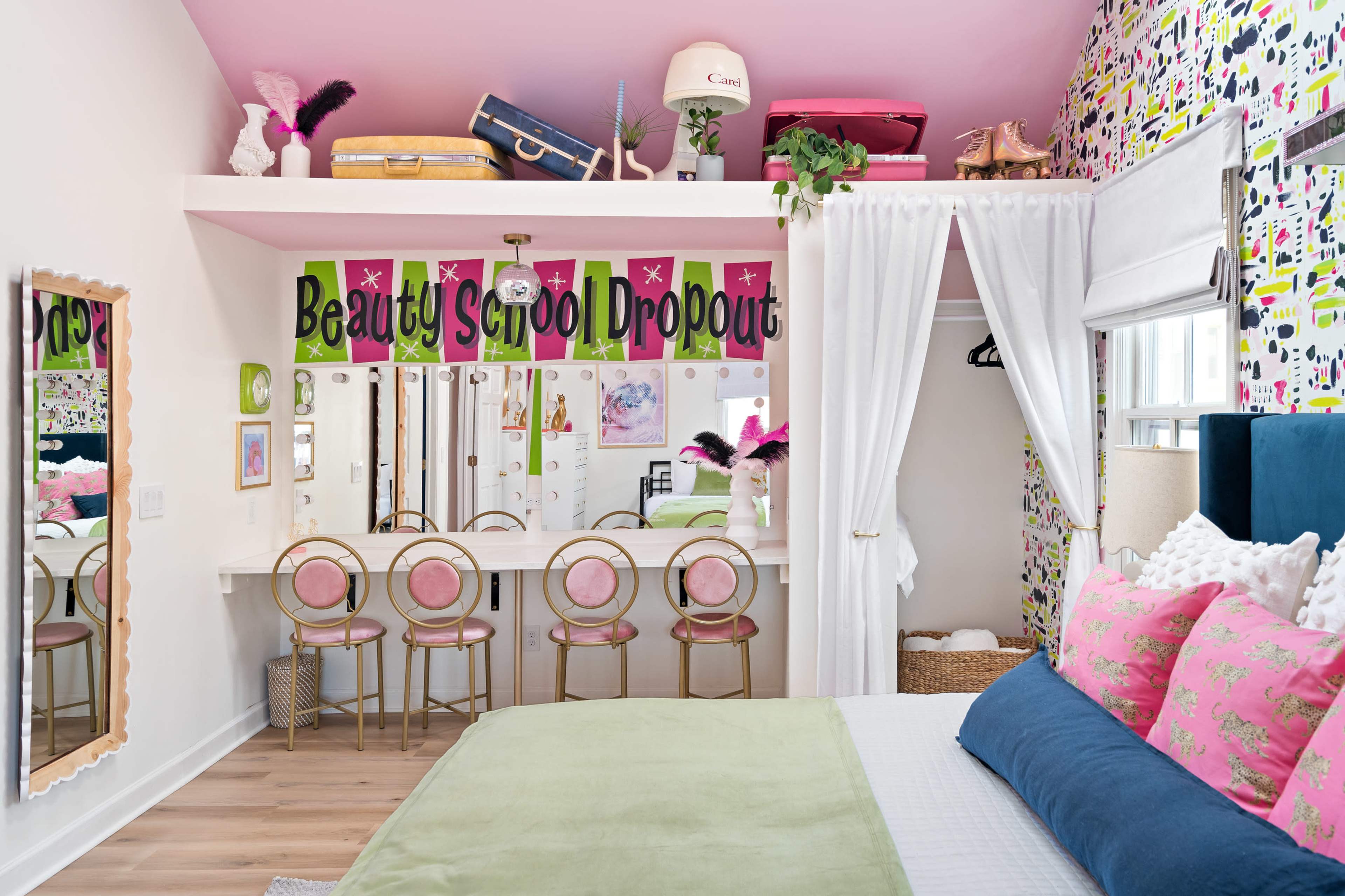 A colorful bedroom featuring a bed, a vanity with pink stools, and a playful, patterned wall adorned with the words "Beauty School Dropout."