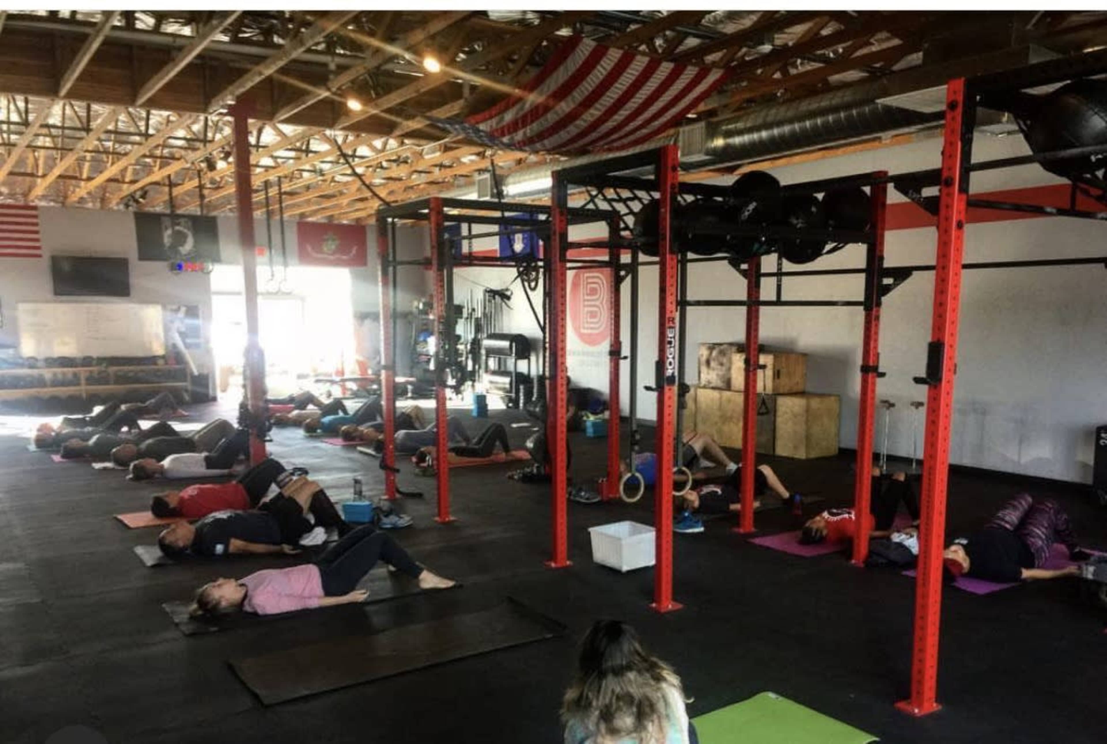 A group of people are lying on yoga mats in a gym, with fitness equipment and an American flag visible in the background.