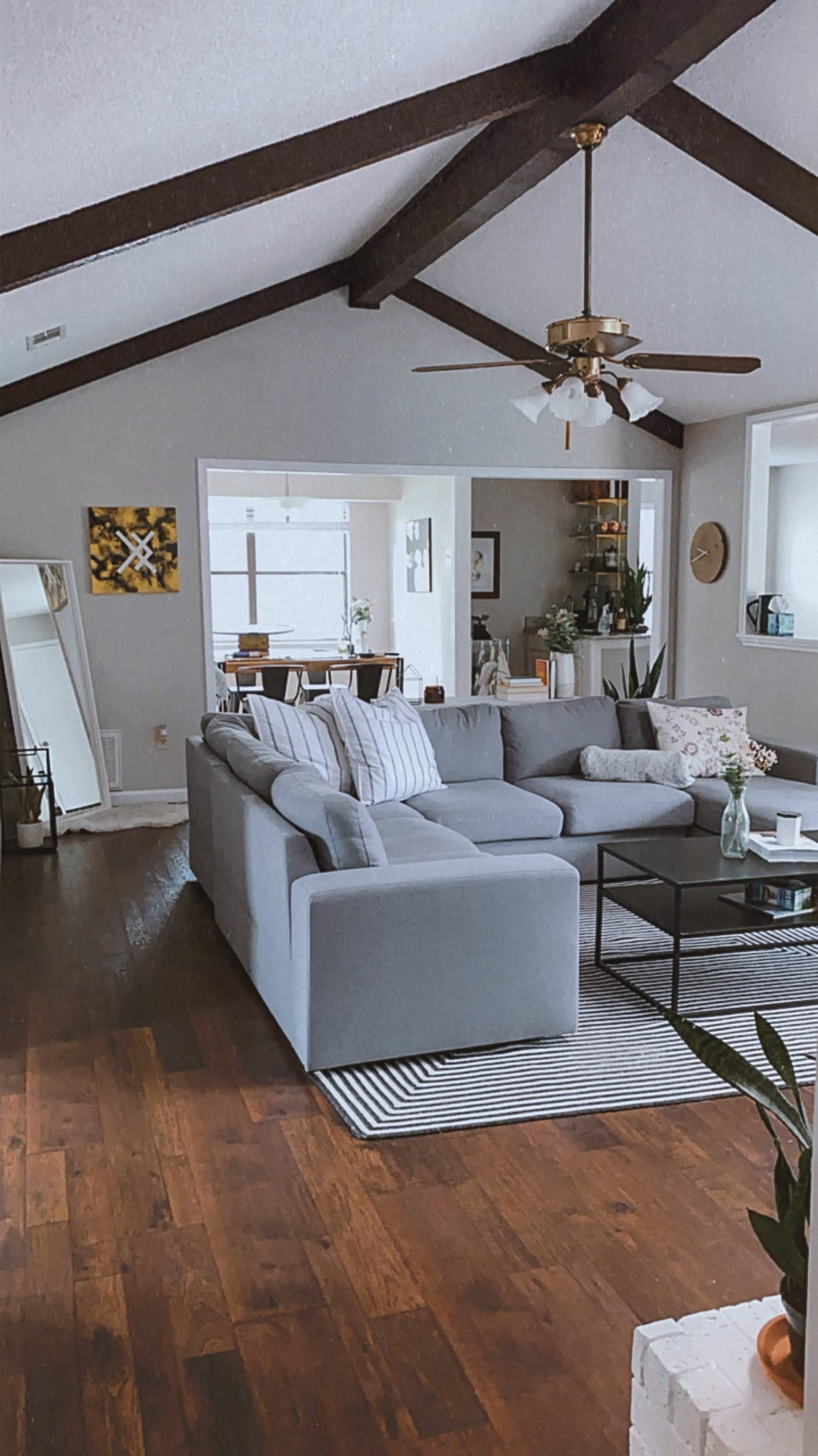 The living room features a gray sectional sofa, wooden beams on the ceiling, and a striped area rug on a hardwood floor.