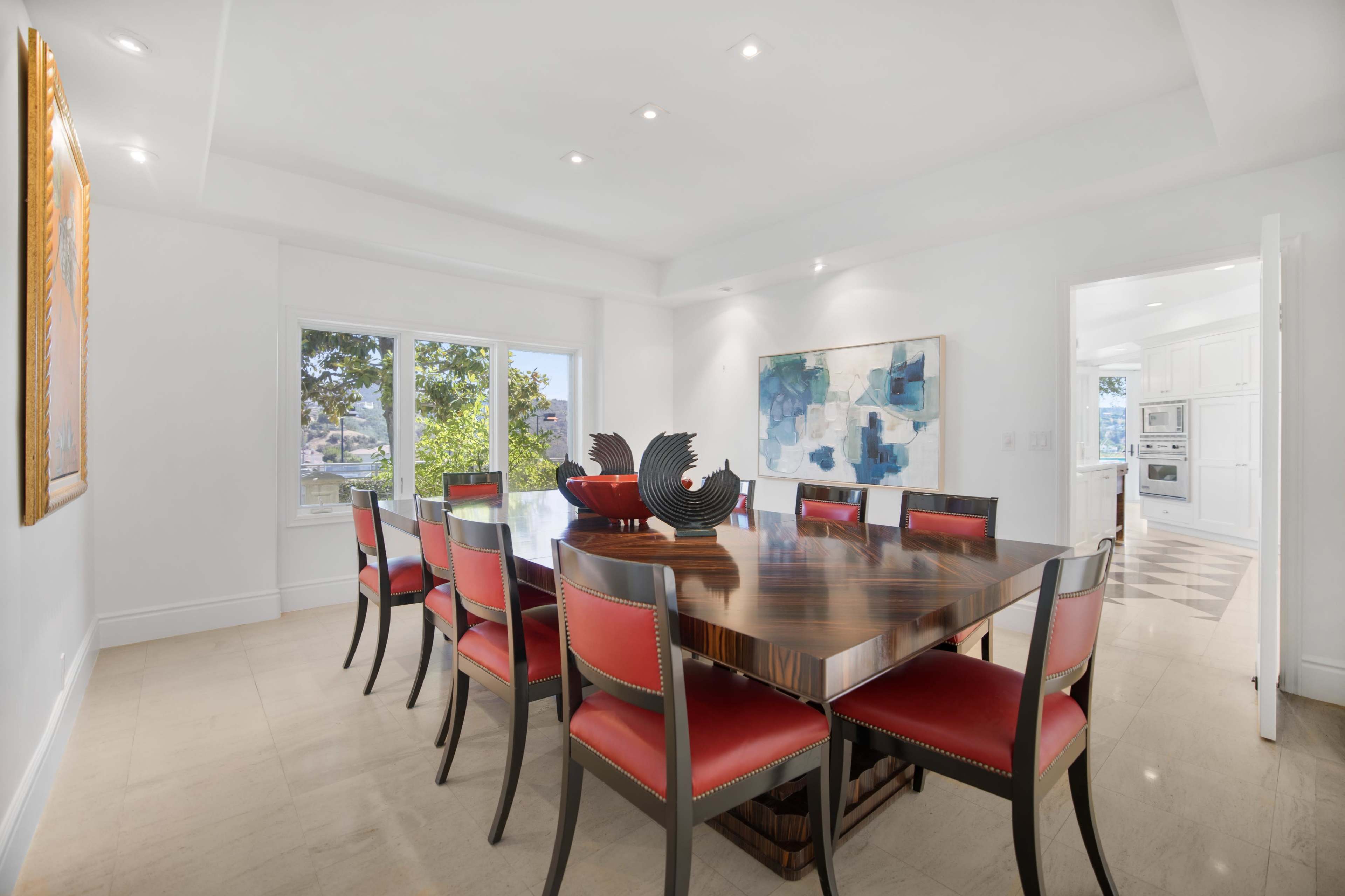 A modern dining room features a large wooden table surrounded by red-upholstered chairs, with artwork on the walls and windows letting in natural light.