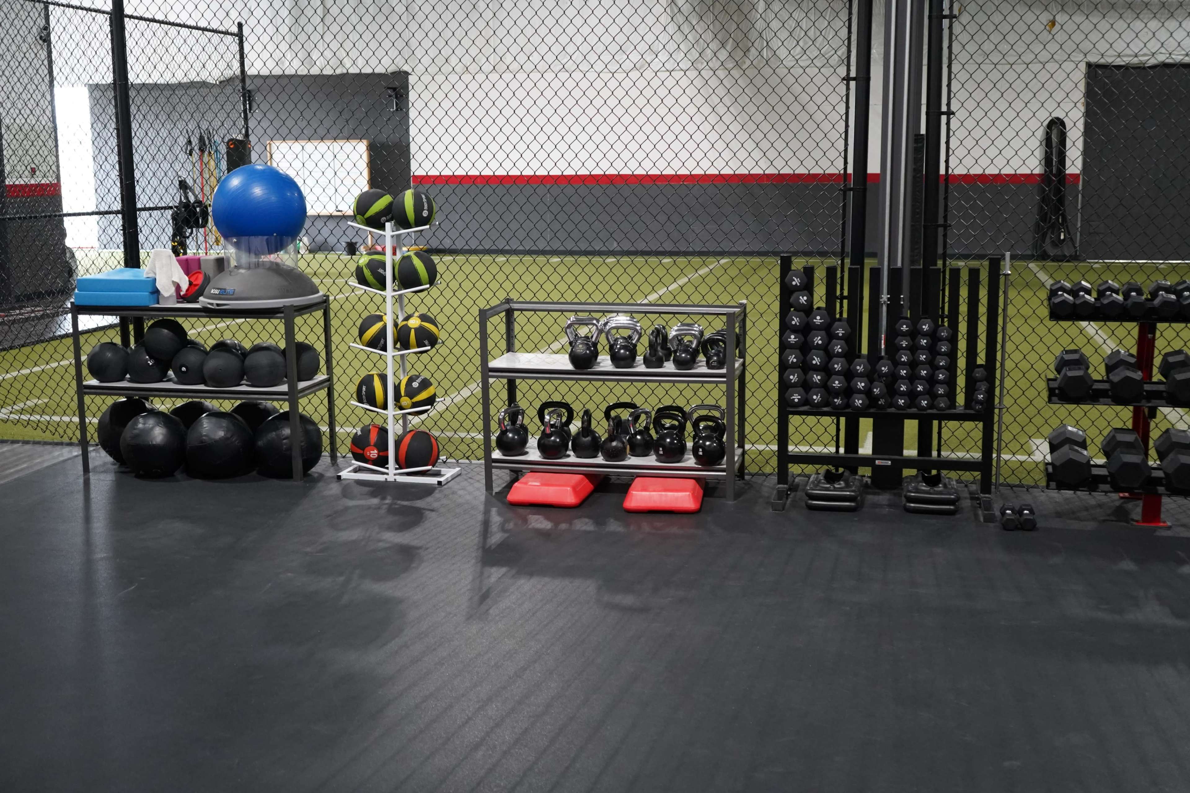 The image shows a gym area with various fitness equipment, including kettlebells, medicine balls, and a stability ball, arranged along a wall and on shelves.