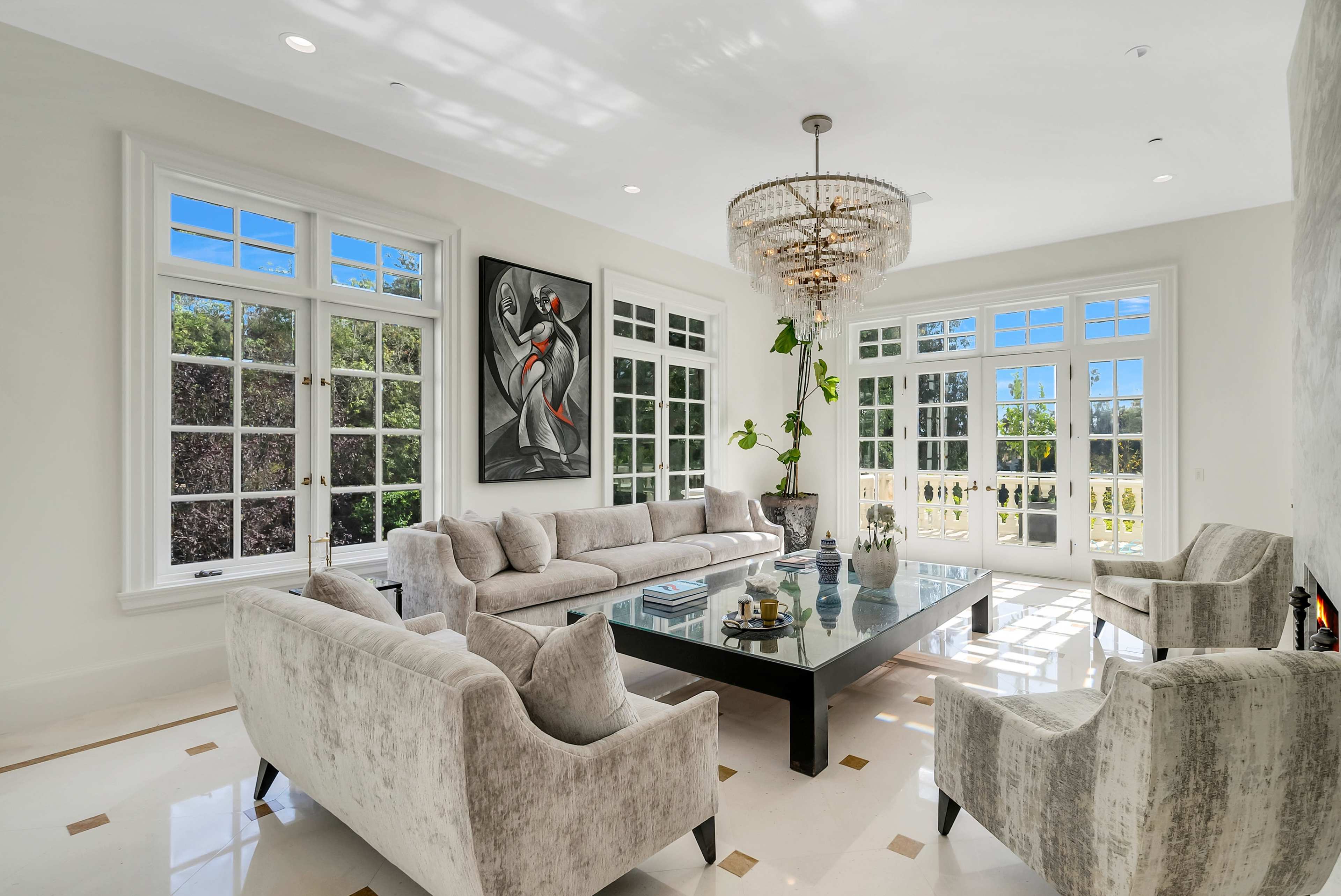 A modern living room features a plush sofa set, a glass coffee table, and large windows allowing natural light to illuminate the space, complemented by a piece of artwork on the wall.