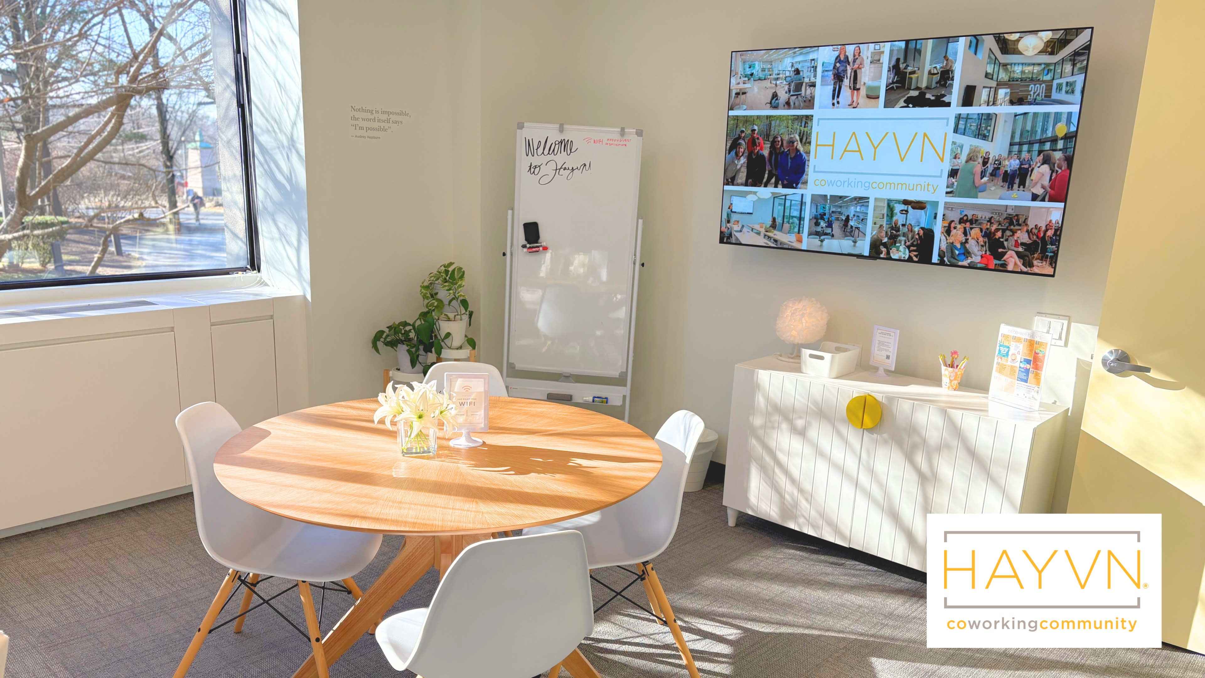 A bright and modern coworking space features a round wooden table surrounded by white chairs, with a large screen displaying community pictures on the wall.
