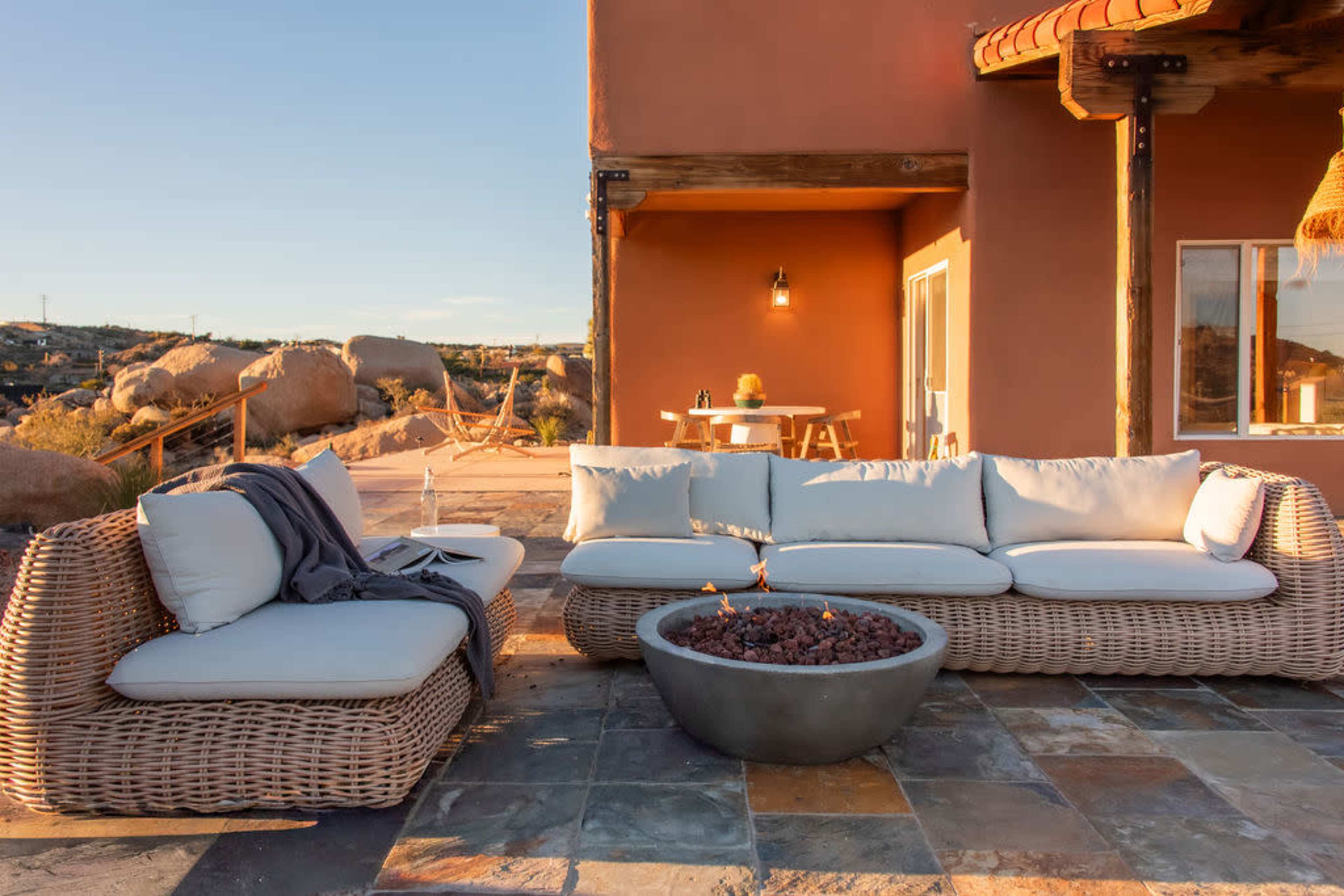The image shows a modern outdoor lounge area with a circular fire pit surrounded by a sectional sofa, set against a backdrop of a desert landscape and a warm-colored building.