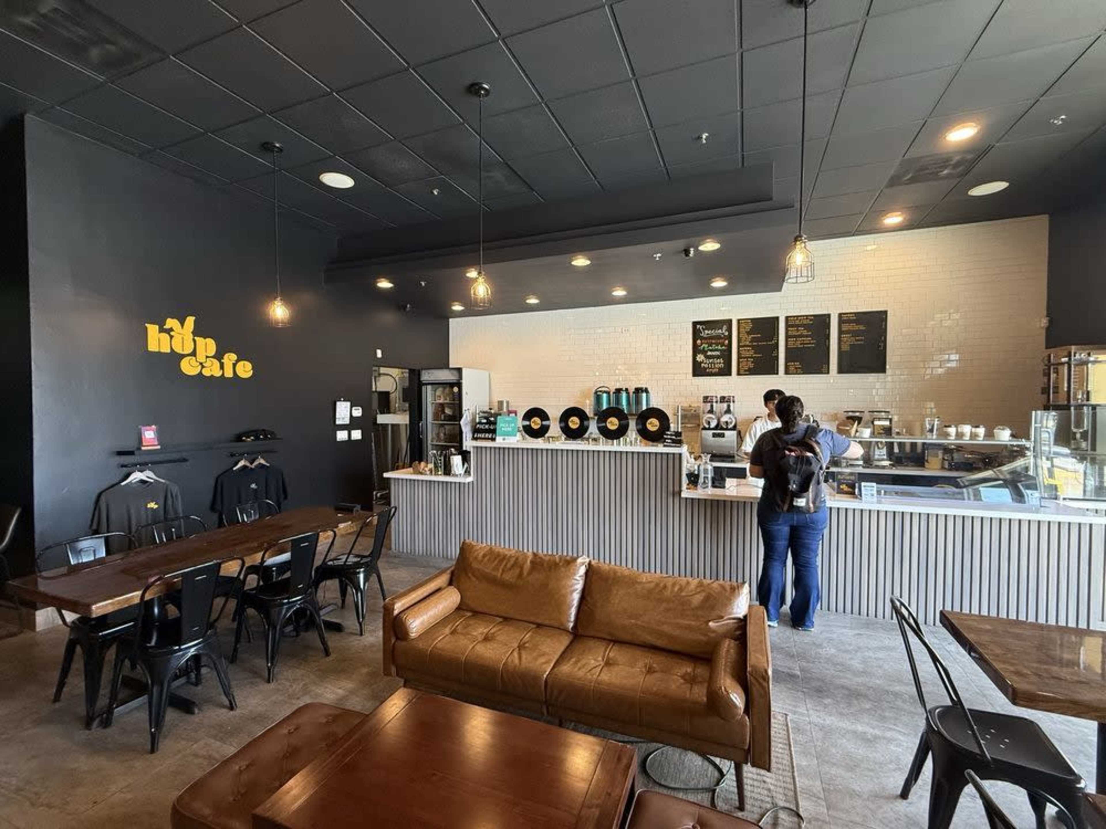 The image shows the interior of a cafe featuring a counter with coffee equipment, a display case, and several tables and chairs in a contemporary setting.