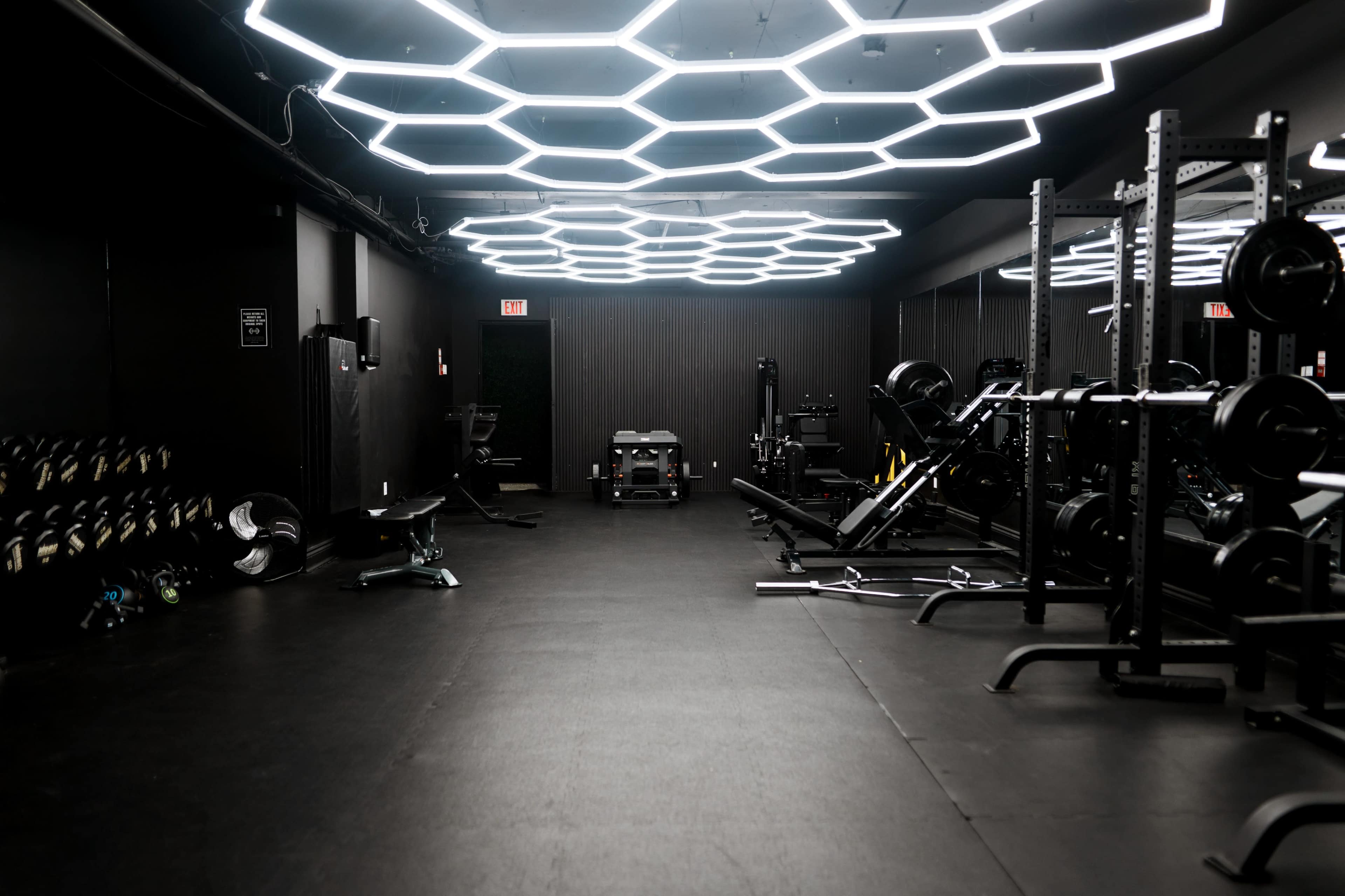 The image shows a gym with a black interior featuring hexagonal ceiling lights, weightlifting equipment, and a neatly organized space.