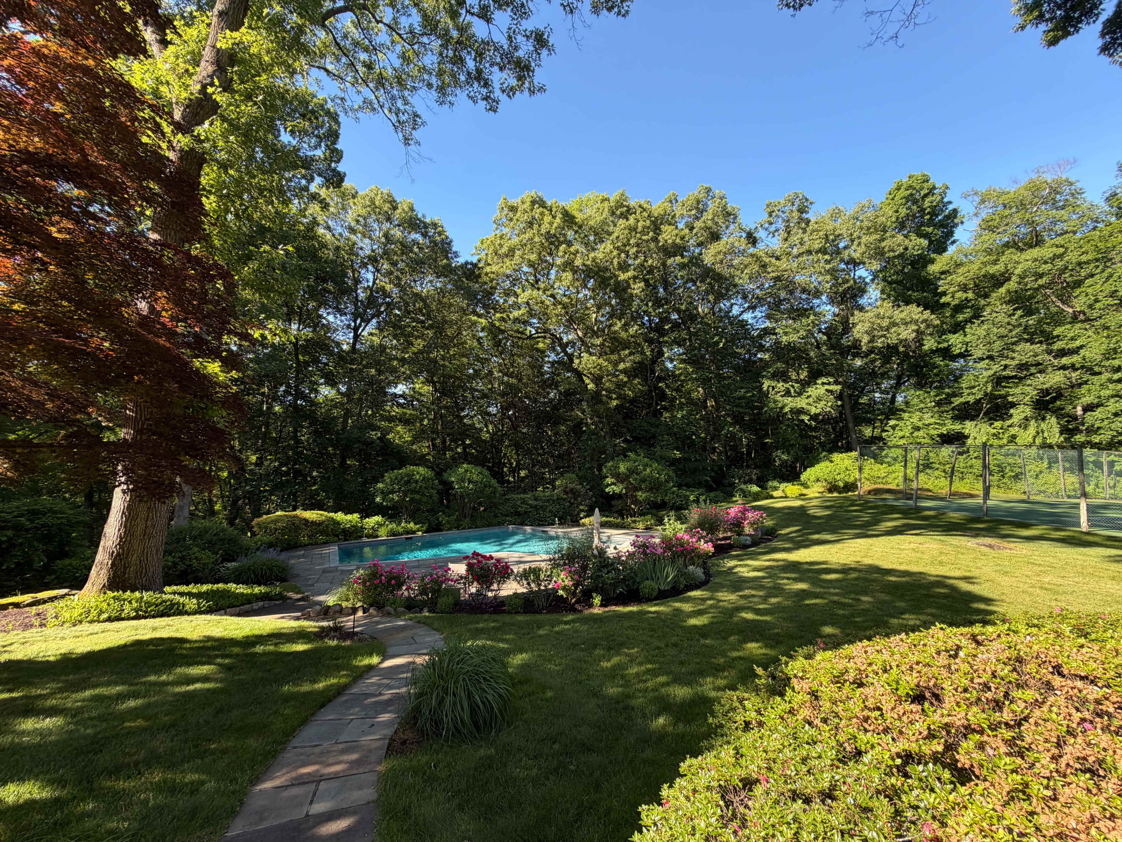 A landscaped backyard features a swimming pool surrounded by vibrant flowers and greenery, with a stone pathway leading through the well-maintained lawn.
