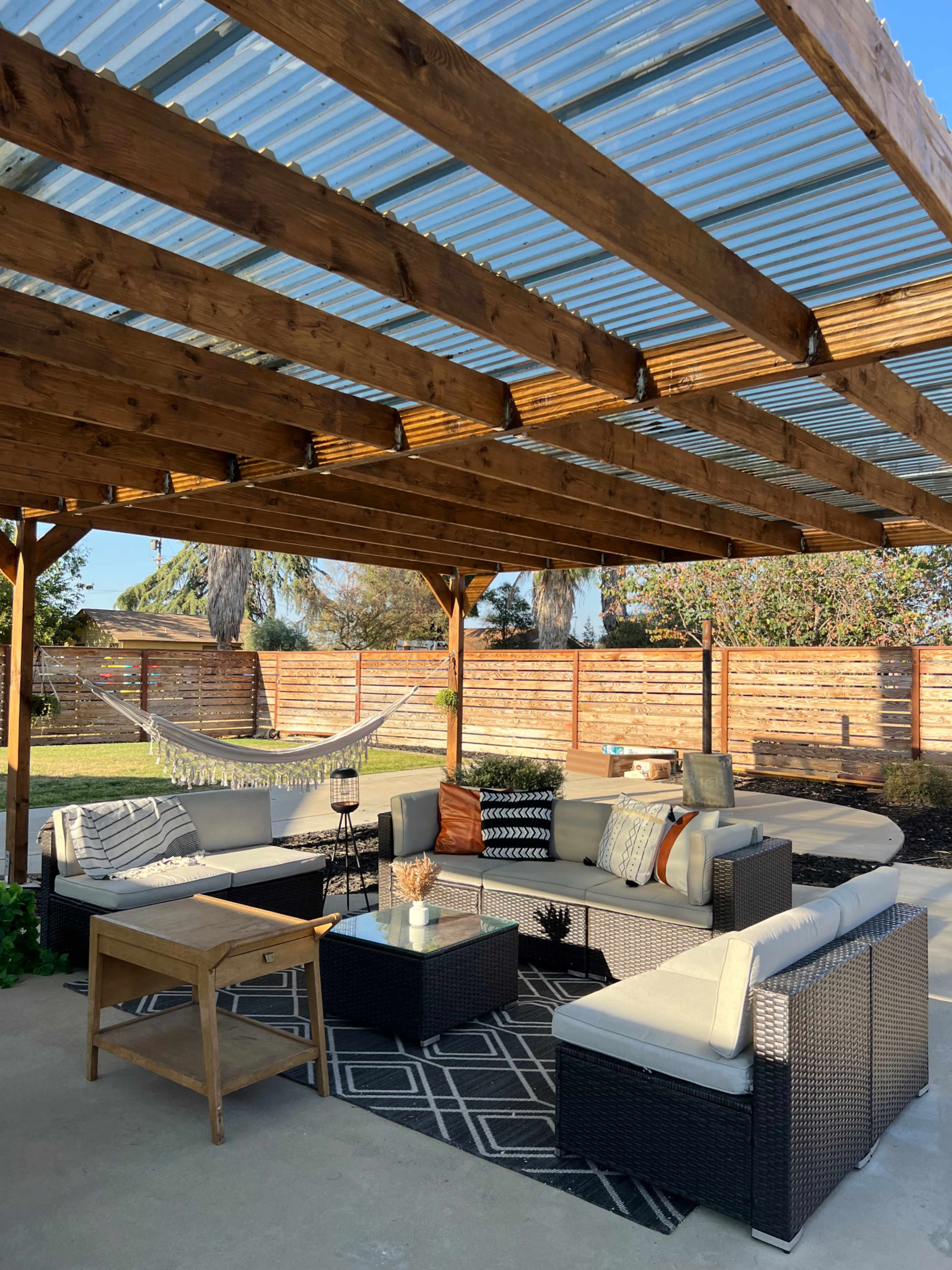 A covered outdoor seating area features a sectional sofa, a coffee table, and a hammock surrounded by a green yard and wooden fencing.