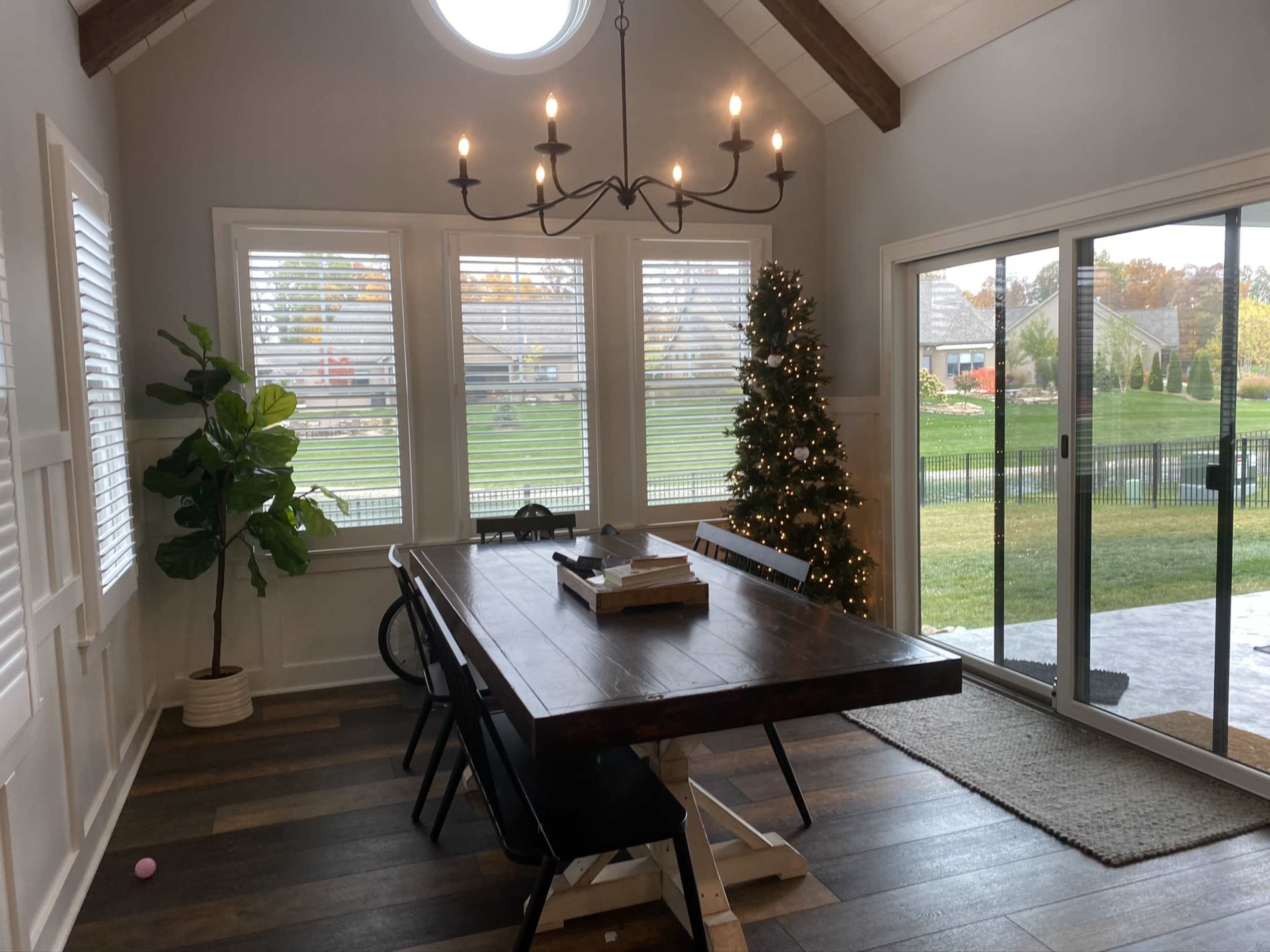 A dining area with a large wooden table, black chairs, a decorative Christmas tree in the corner, and large windows letting in natural light.