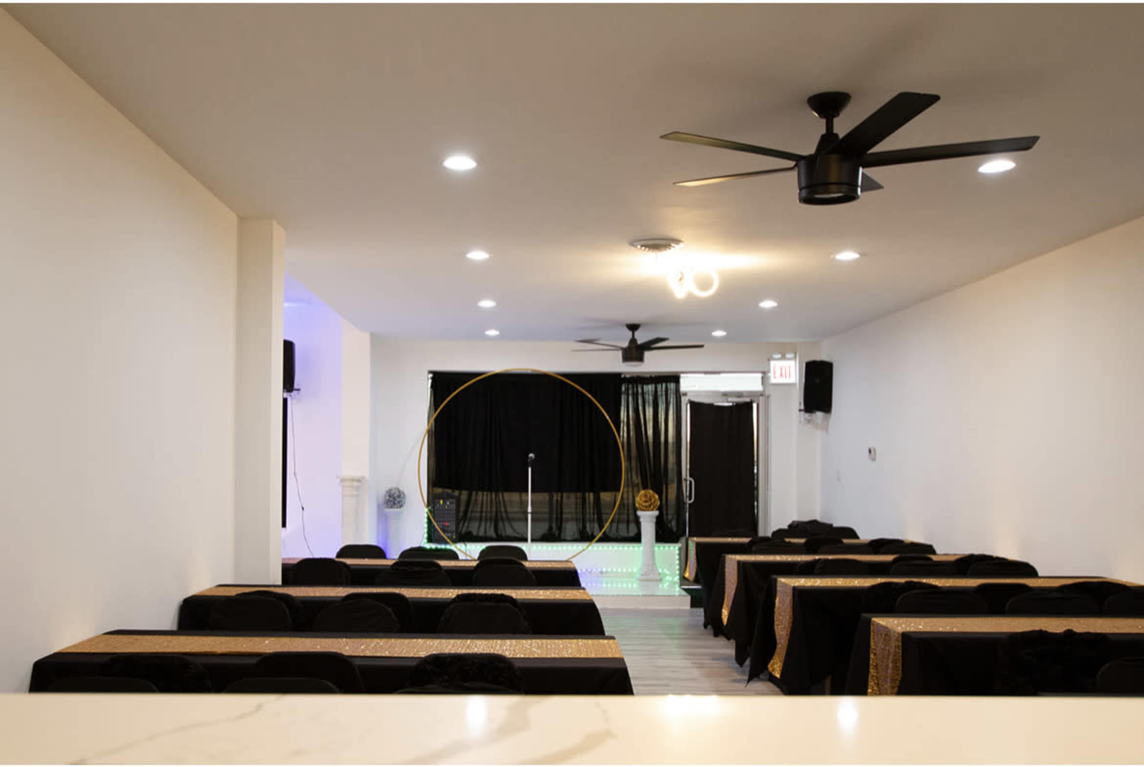 The image shows a spacious event room with rows of tables covered in black cloth and gold table runners, a microphone stand at the front, and black curtains in the background.