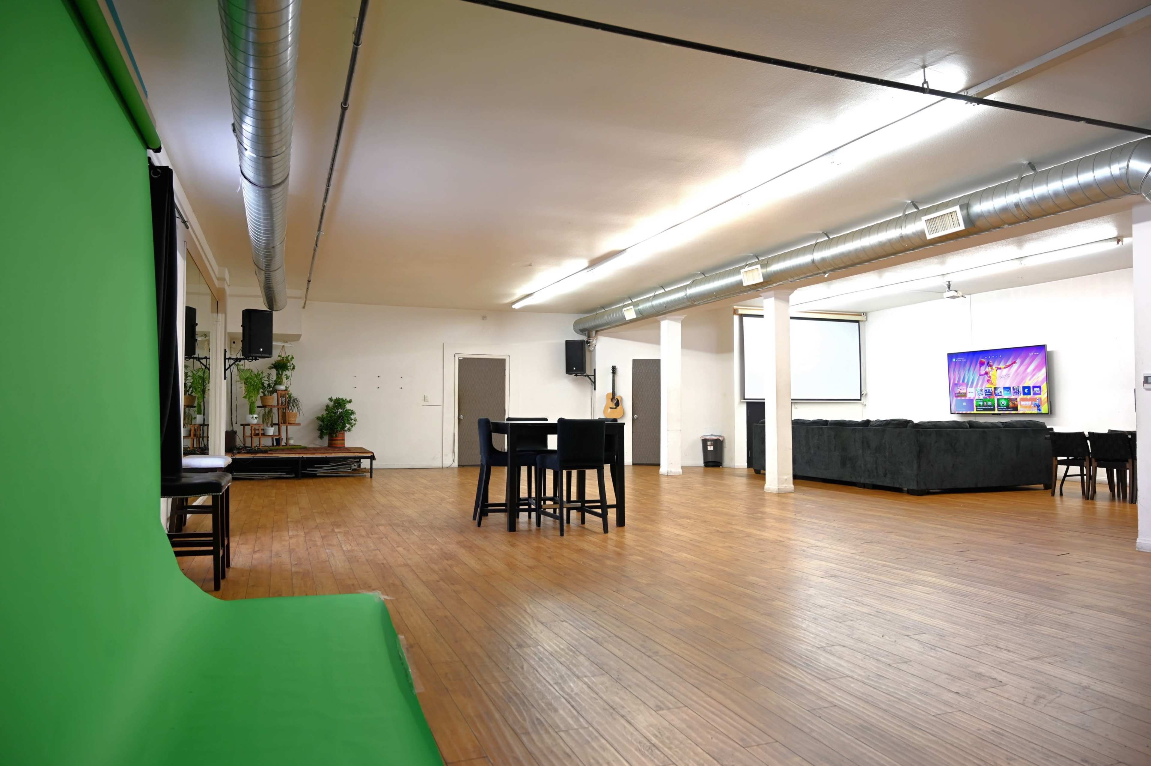 A spacious, empty room with wooden floors, featuring a green backdrop on the left, seating areas, and a projector screen at the far end.