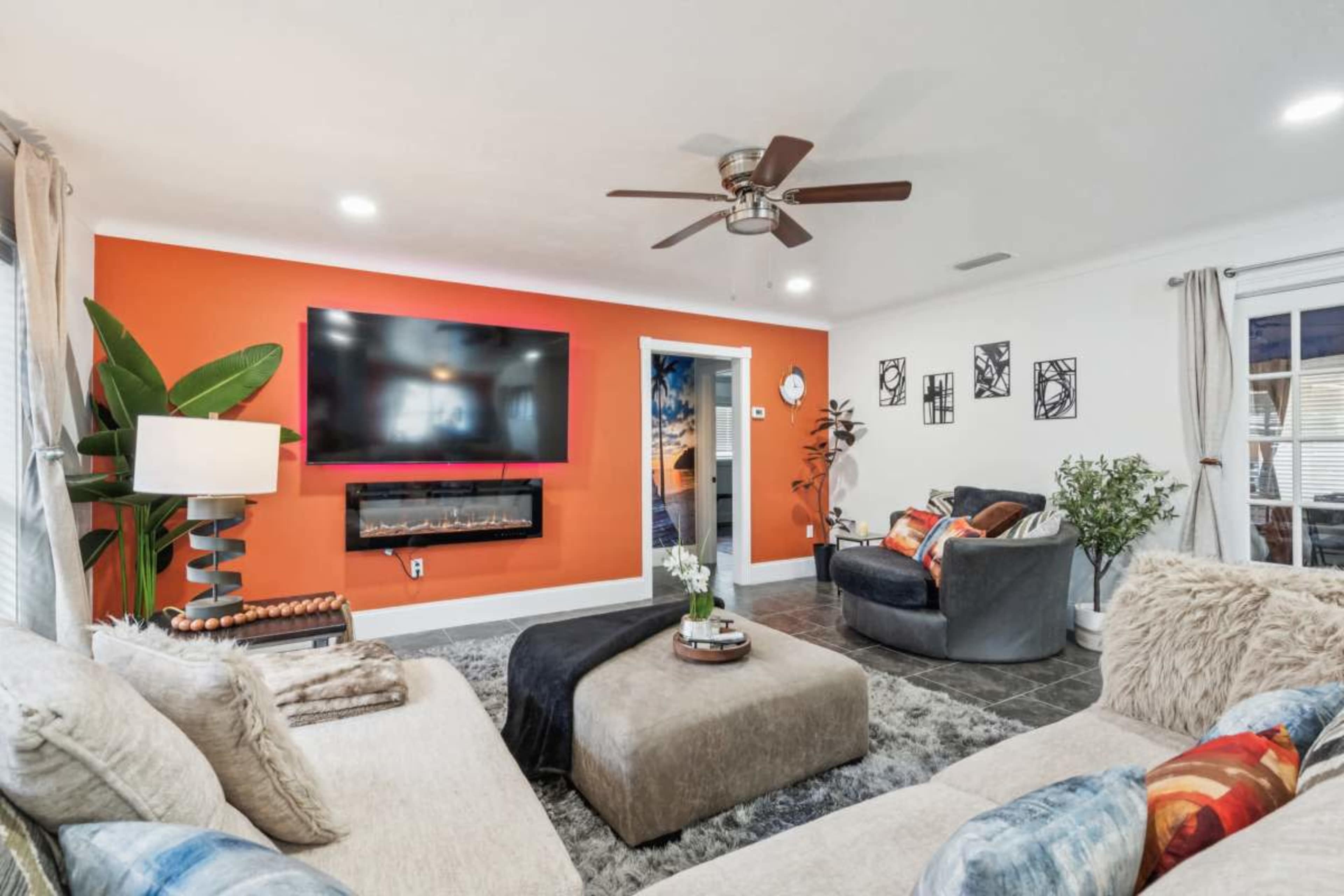 The living room features a large sectional sofa, a mounted television on an orange accent wall, and a modern electric fireplace beneath it.