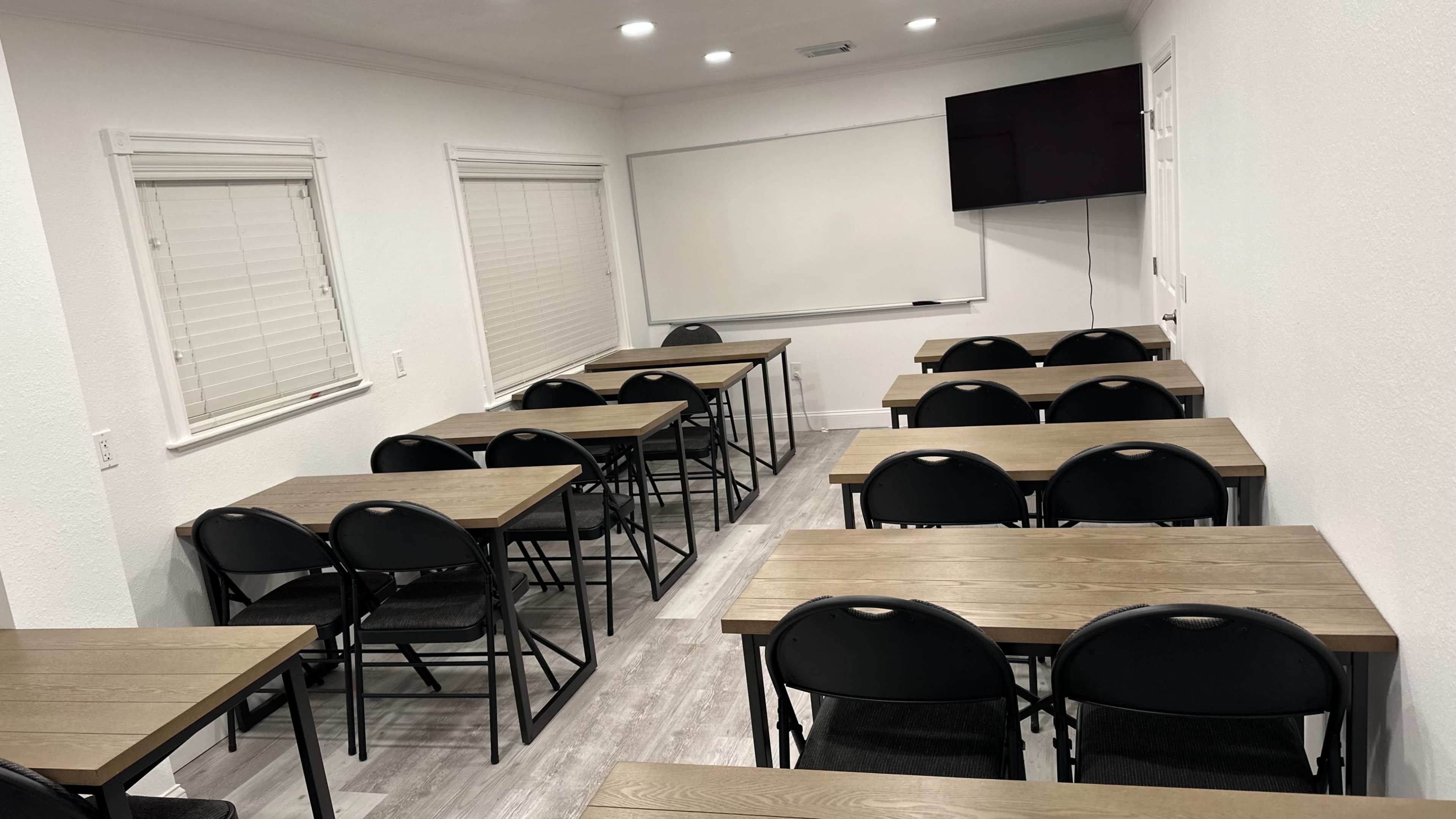 The image shows a classroom with several rows of tables and chairs arranged facing a whiteboard and a wall-mounted television.