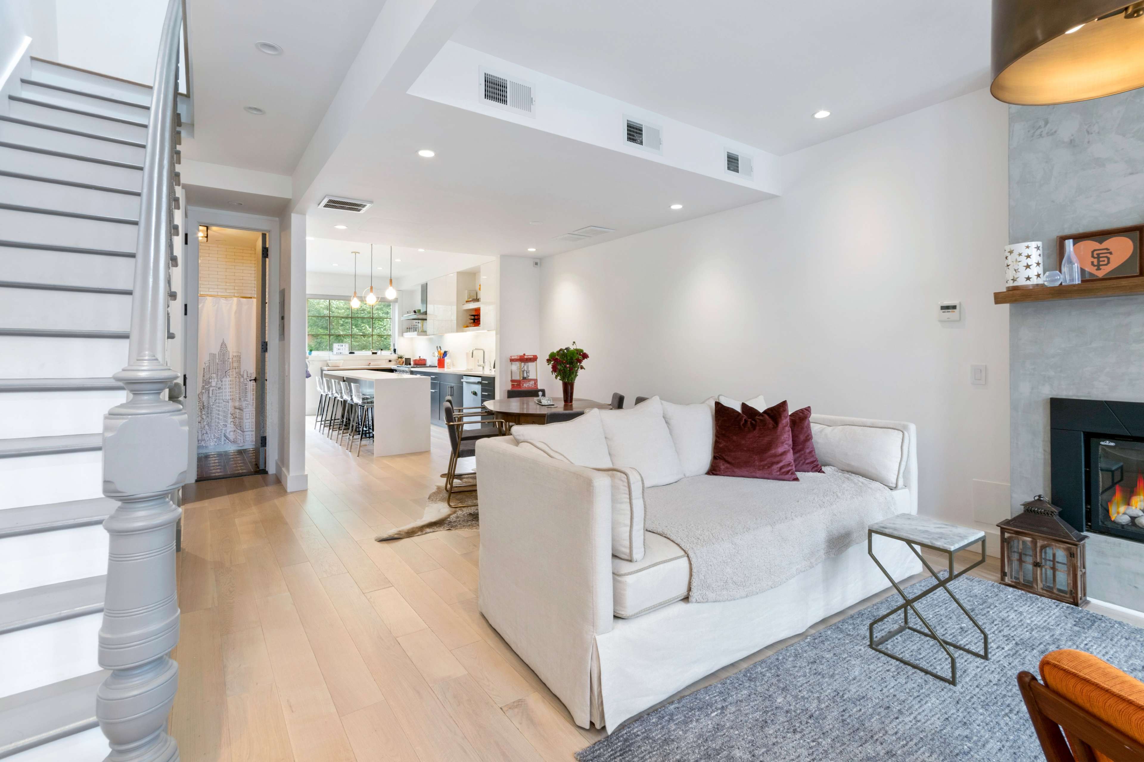 A modern living room with a white sofa, gray rug, and a fireplace, connected to a kitchen area with bar seating visible in the background.
