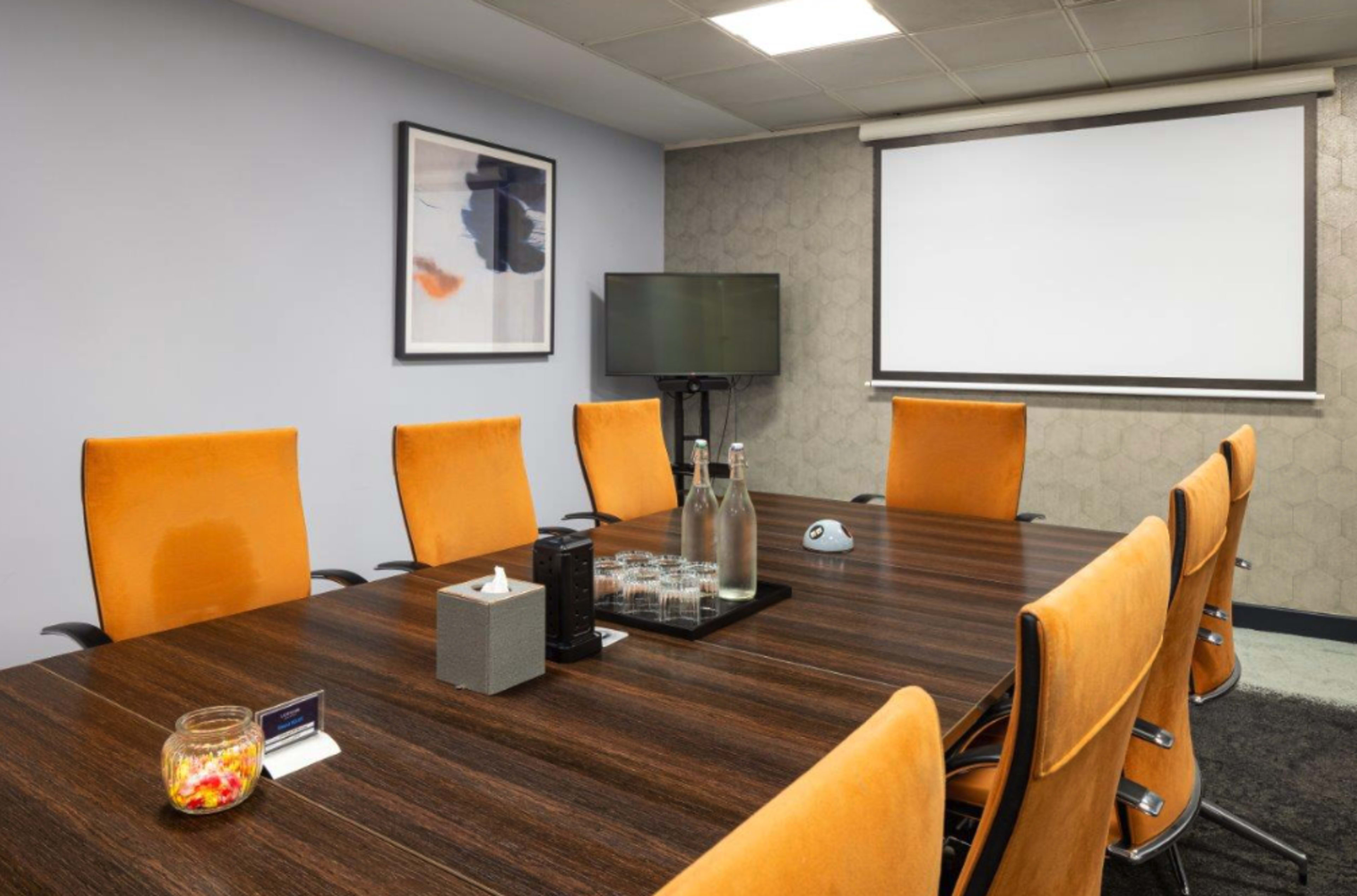 The image shows a conference room with a rectangular wooden table surrounded by orange chairs, a television, and a projection screen.