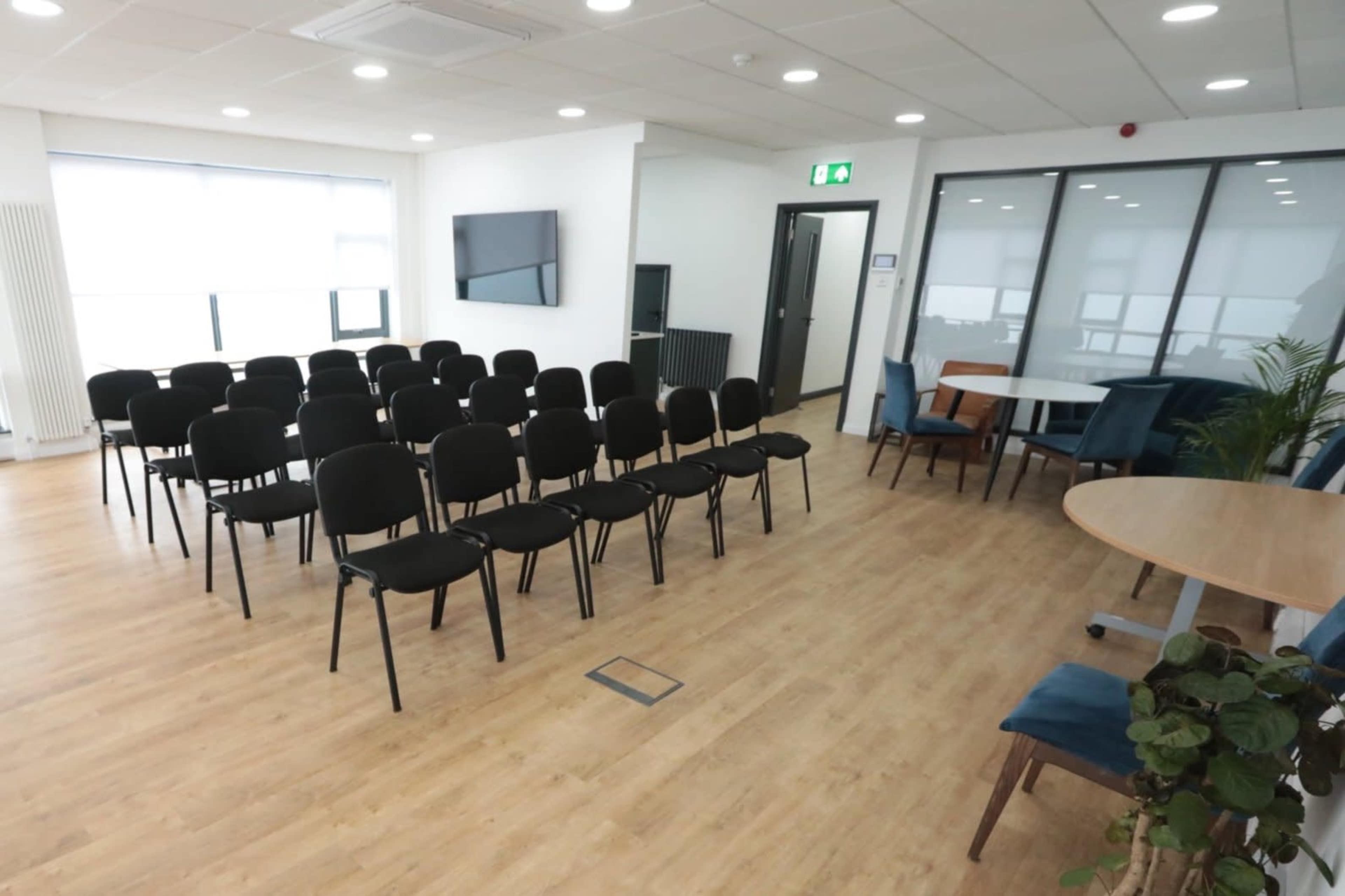 A modern conference room features a row of black chairs arranged for an audience and a separate area with tables and plants.