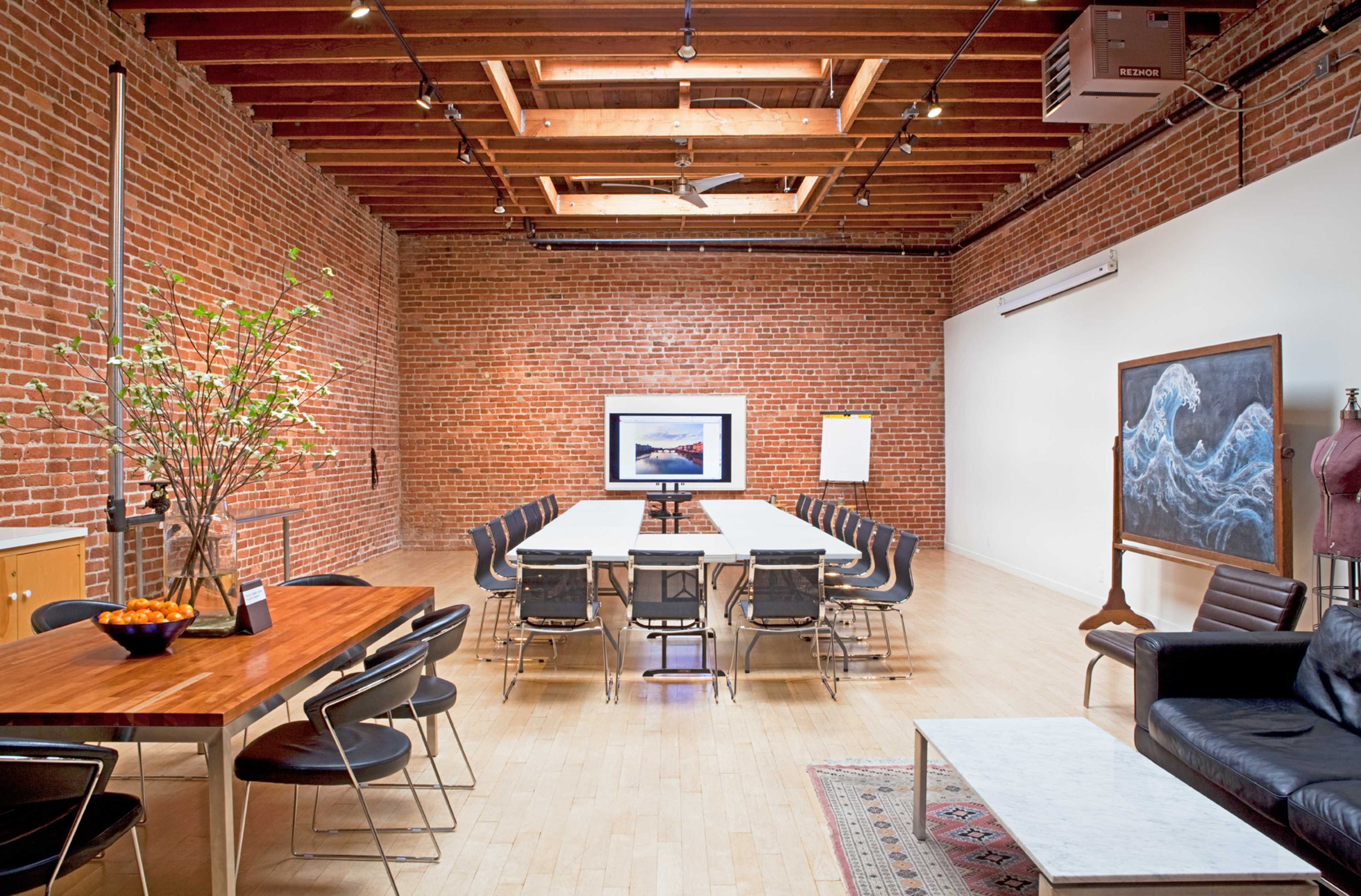 A spacious room with exposed brick walls, a wooden beam ceiling, a long central table surrounded by chairs, and various pieces of art displayed on the walls.