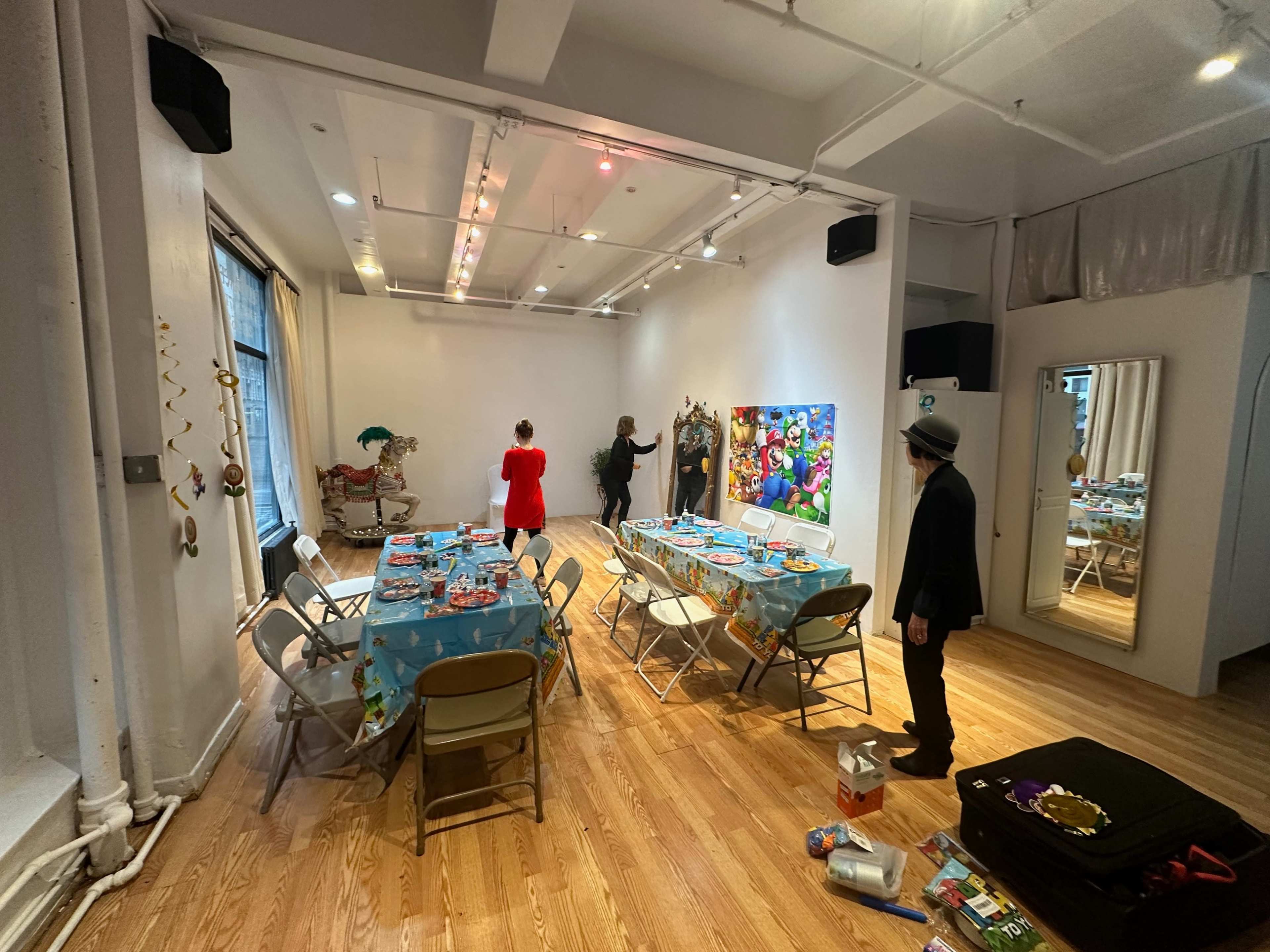 A spacious room is set up for a gathering, featuring tables with blue tablecloths and decorations, while individuals prepare decor on the walls.