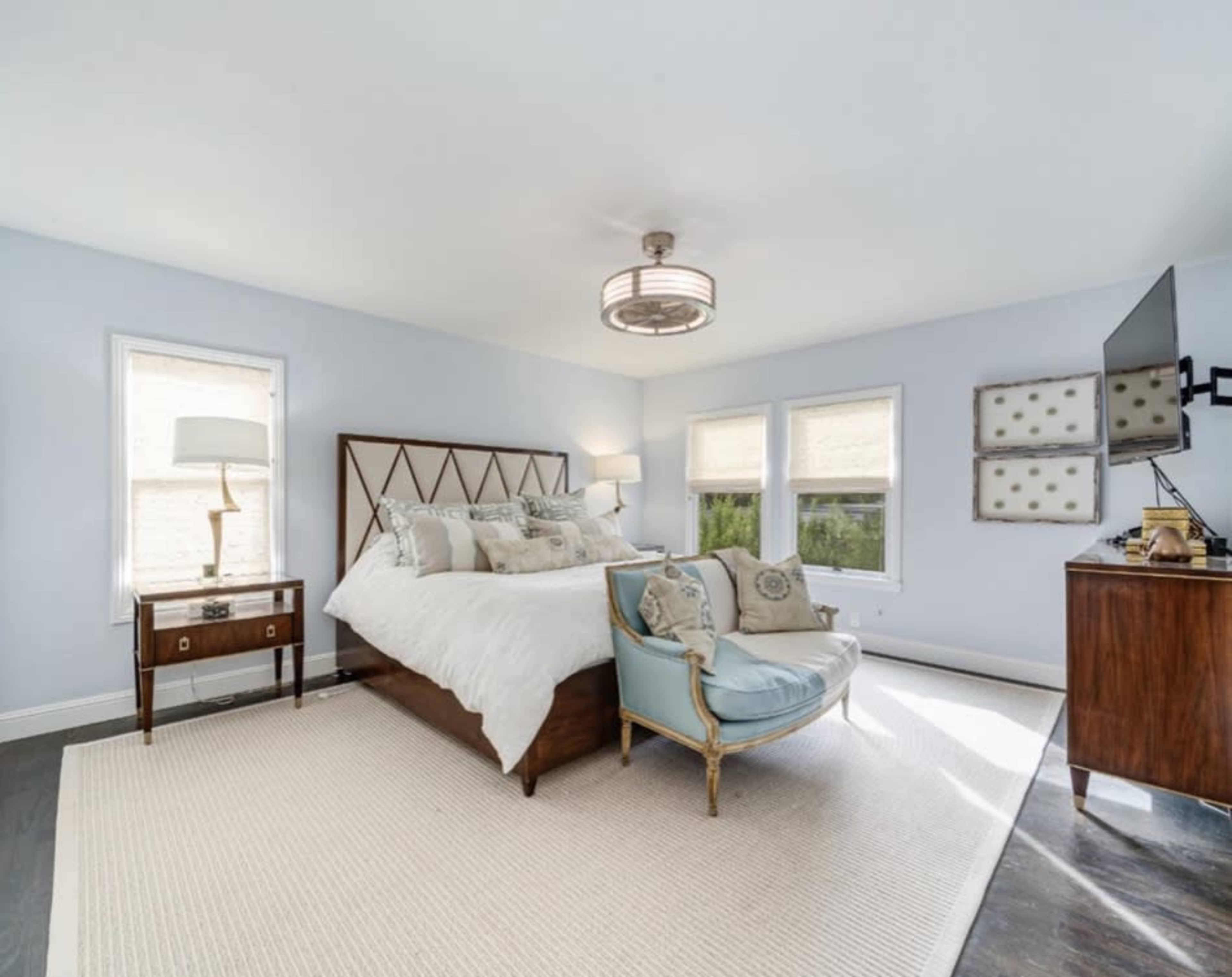 A bedroom featuring a large bed with decorative pillows, a small side table with a lamp, an accent chair, and a television mounted on the wall, all set against light blue walls and a light area rug.