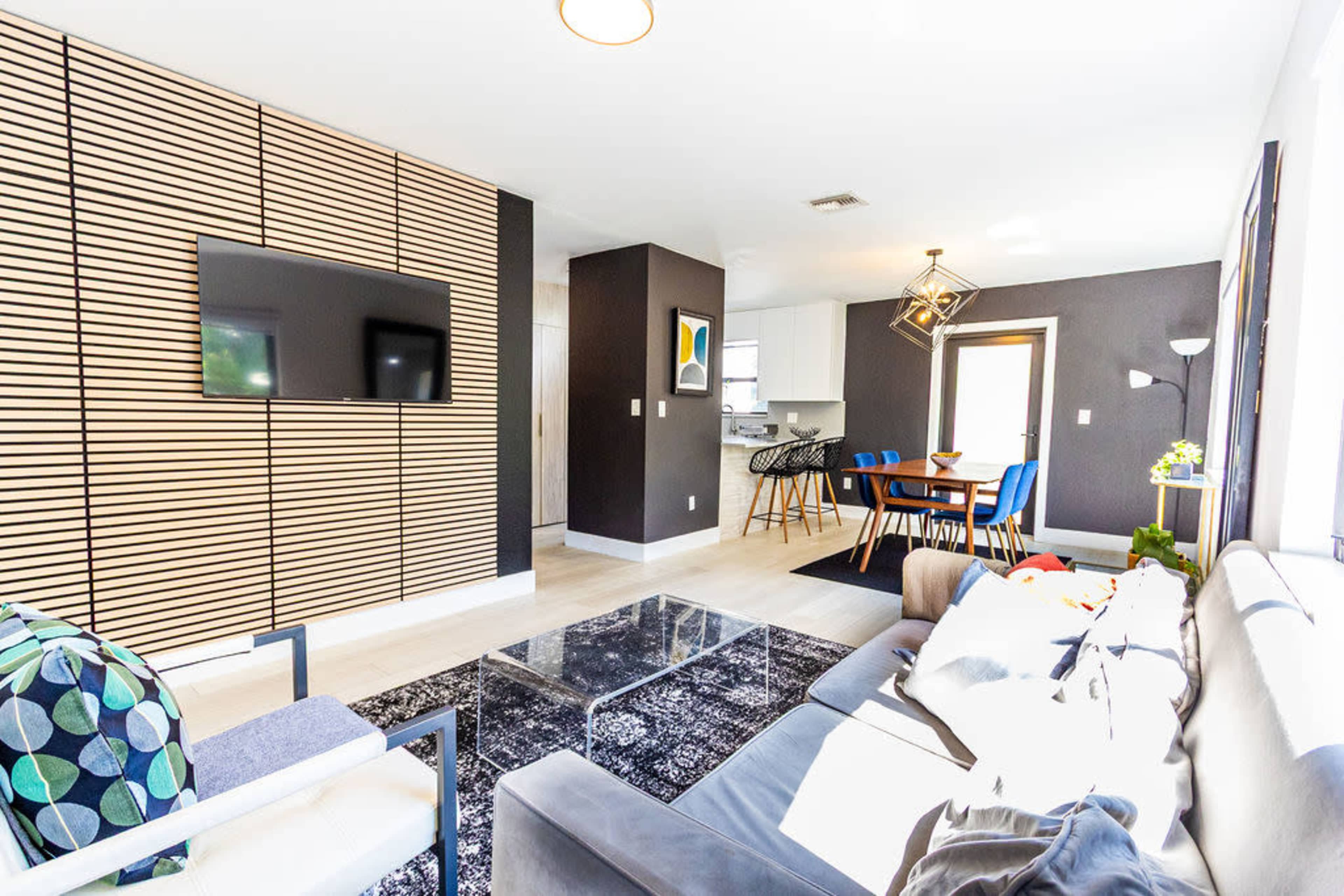 The image shows a modern living room with a sectional sofa, a glass coffee table, a textured wall with a television, and a dining area featuring a wooden table and chairs.