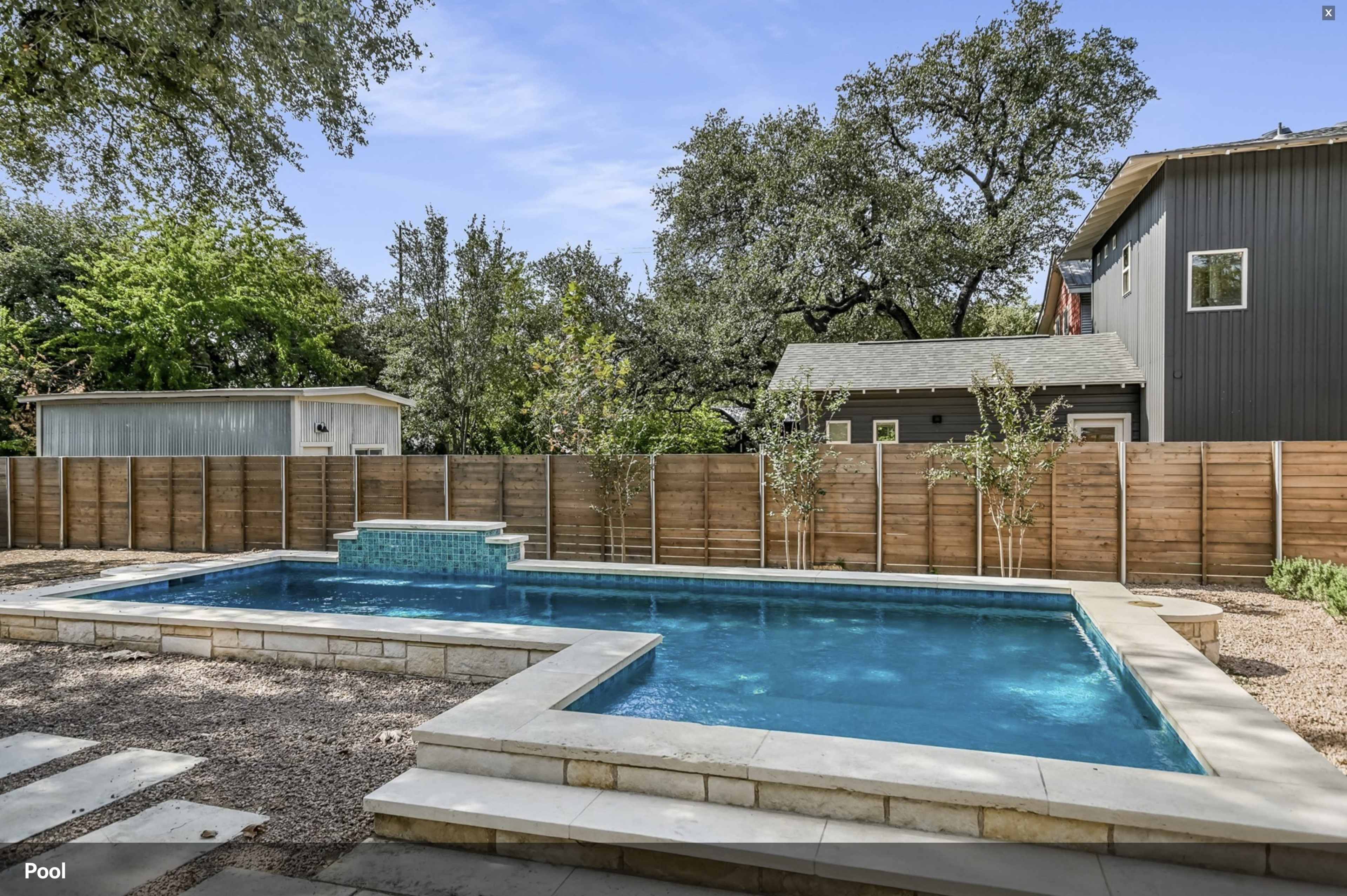 The image shows a backyard with a rectangular pool surrounded by stone decking and bordered by wooden fences, with trees and a house visible in the background.