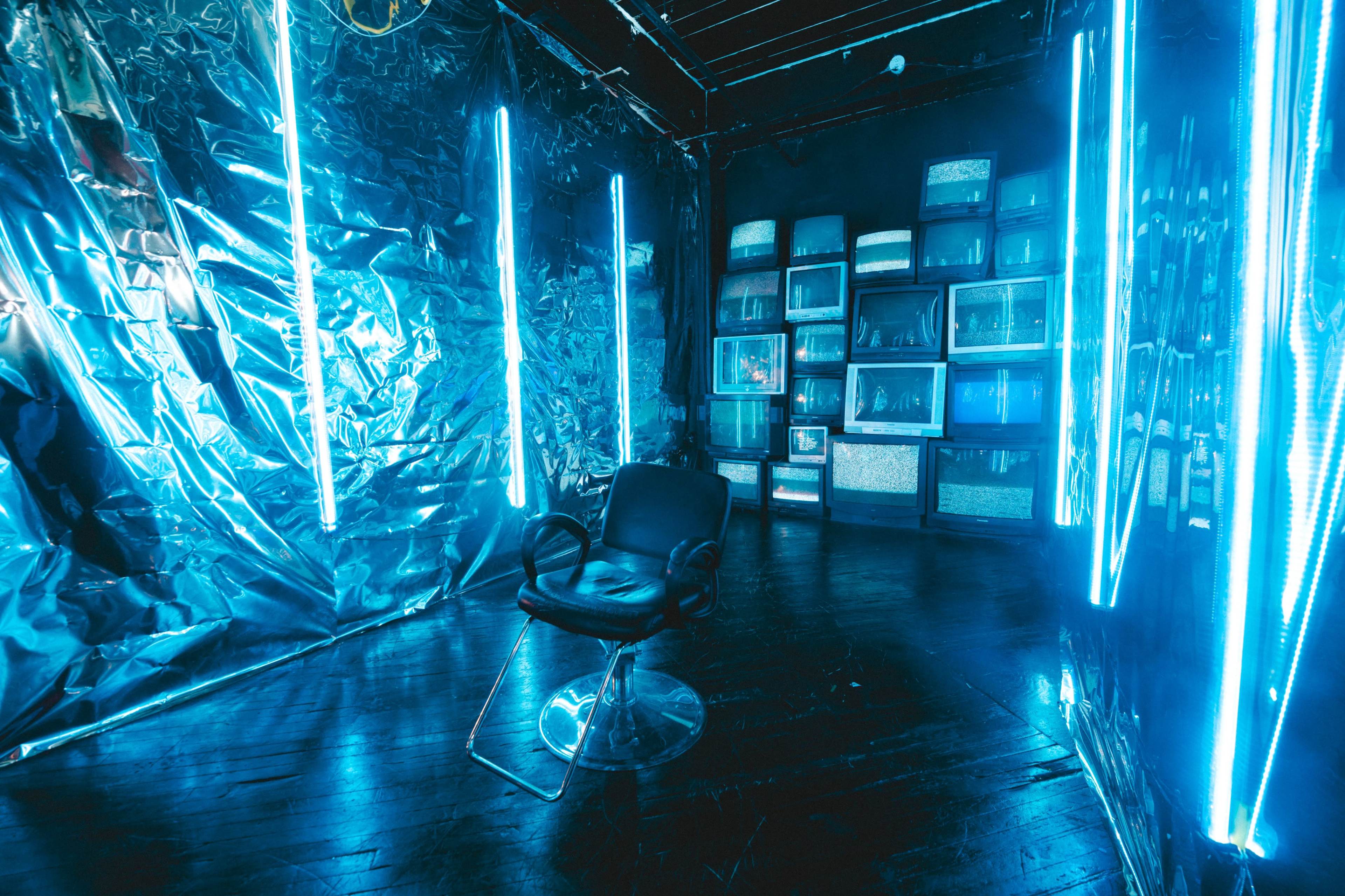 The image shows a room with metallic walls, illuminated by blue neon lights, featuring a black chair and a wall of vintage televisions.