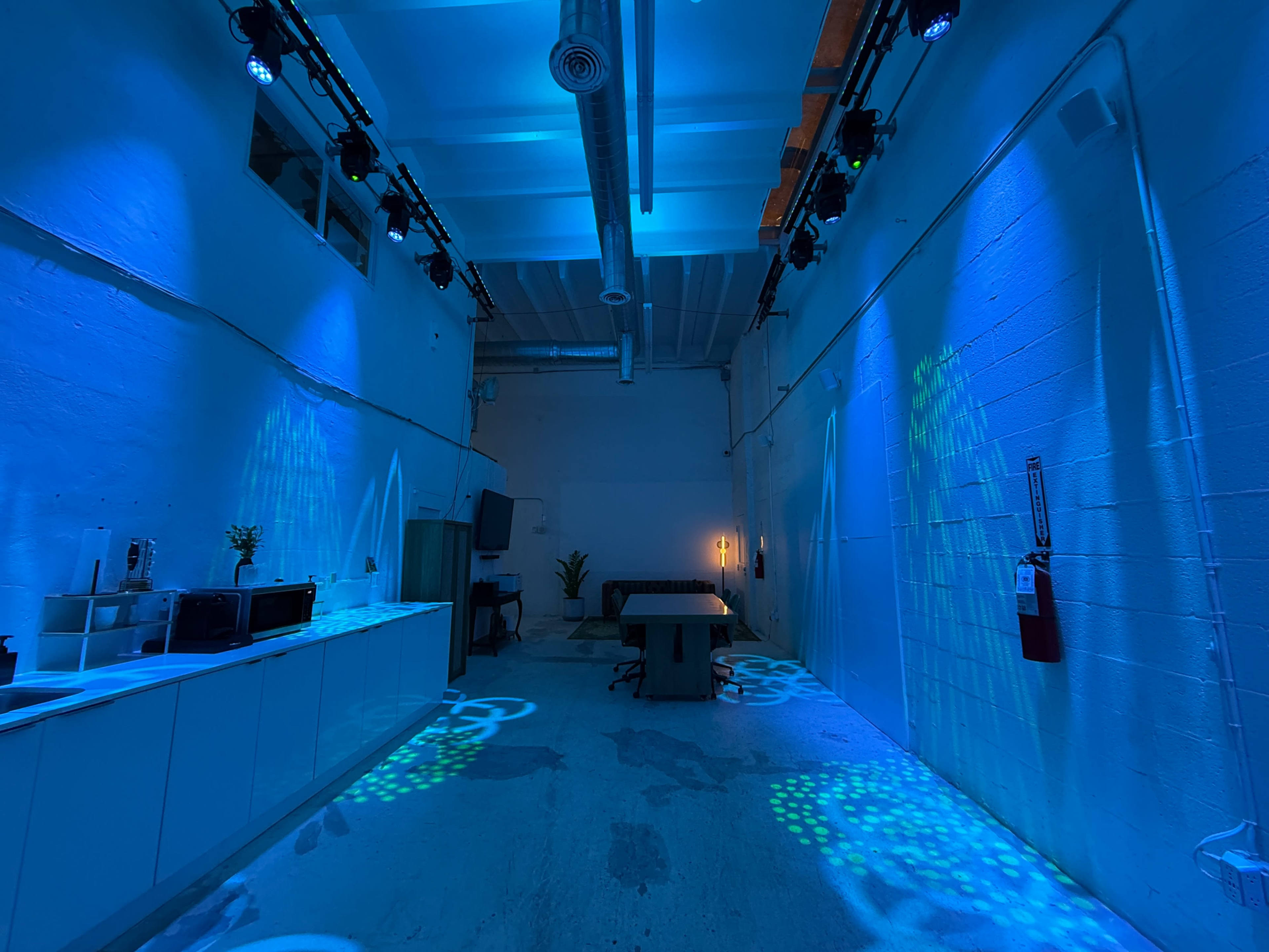 The image shows a spacious, illuminated room with blue lighting, a large table in the center, and kitchen appliances along one wall.