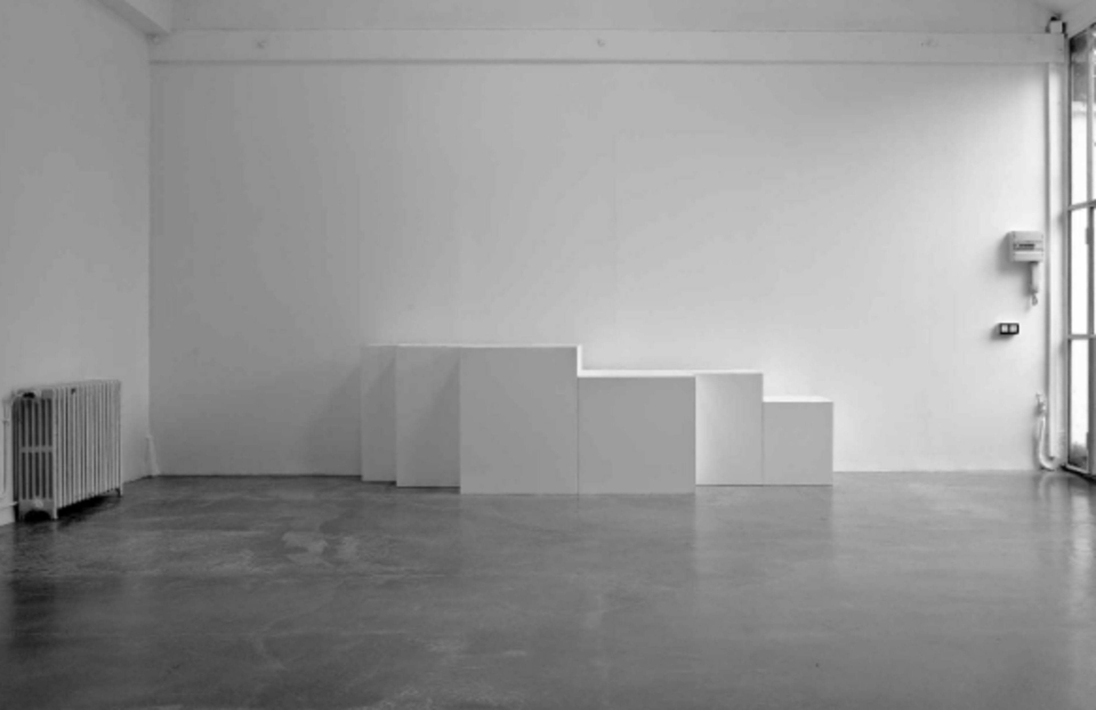 The image depicts a minimalist gallery space featuring a series of white, stepped cuboid structures against a plain white wall.