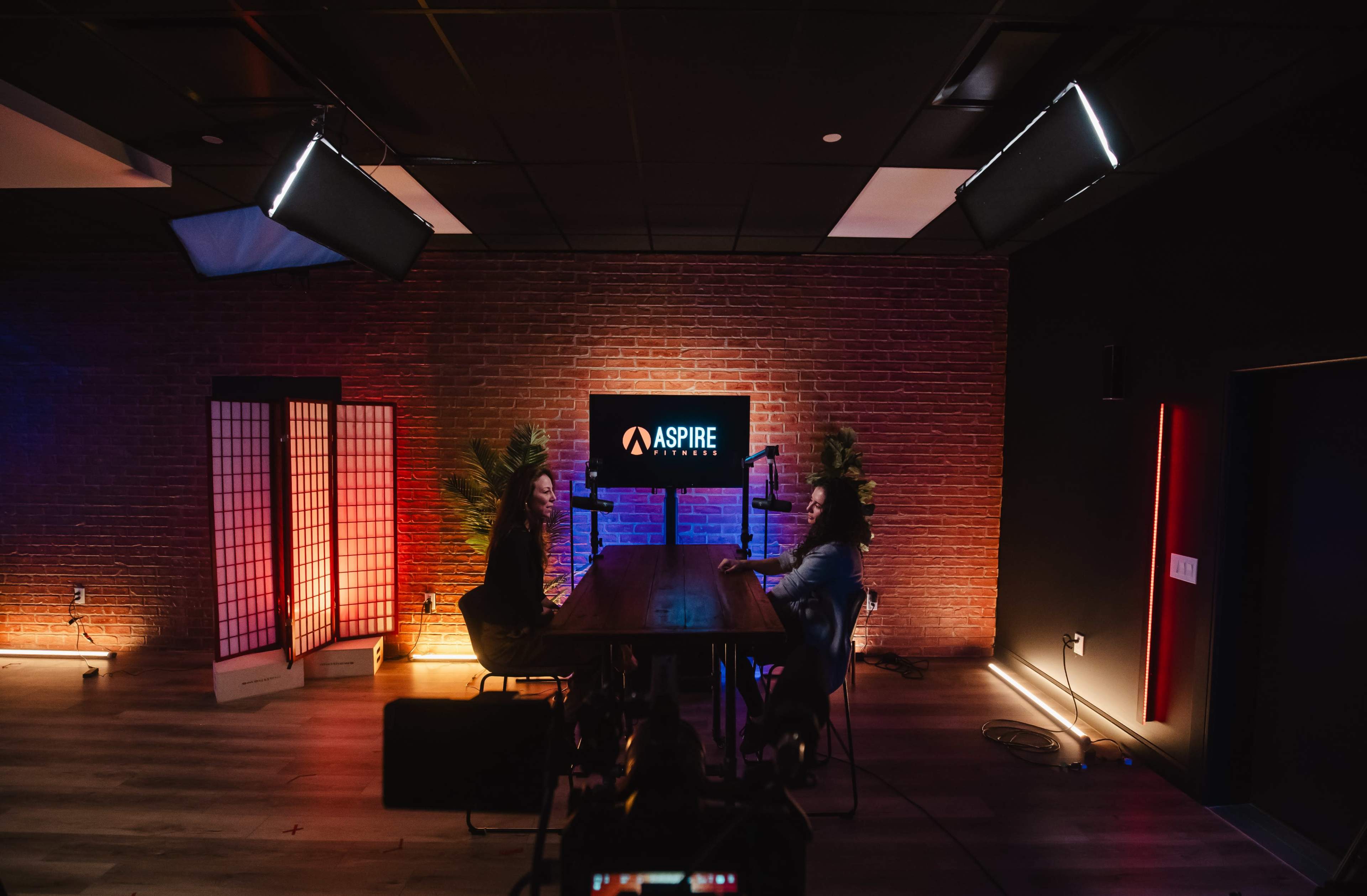 A well-lit studio setting features two individuals seated at a wooden table with microphones, against a backdrop of brick walls and an "ASPIRE" sign.