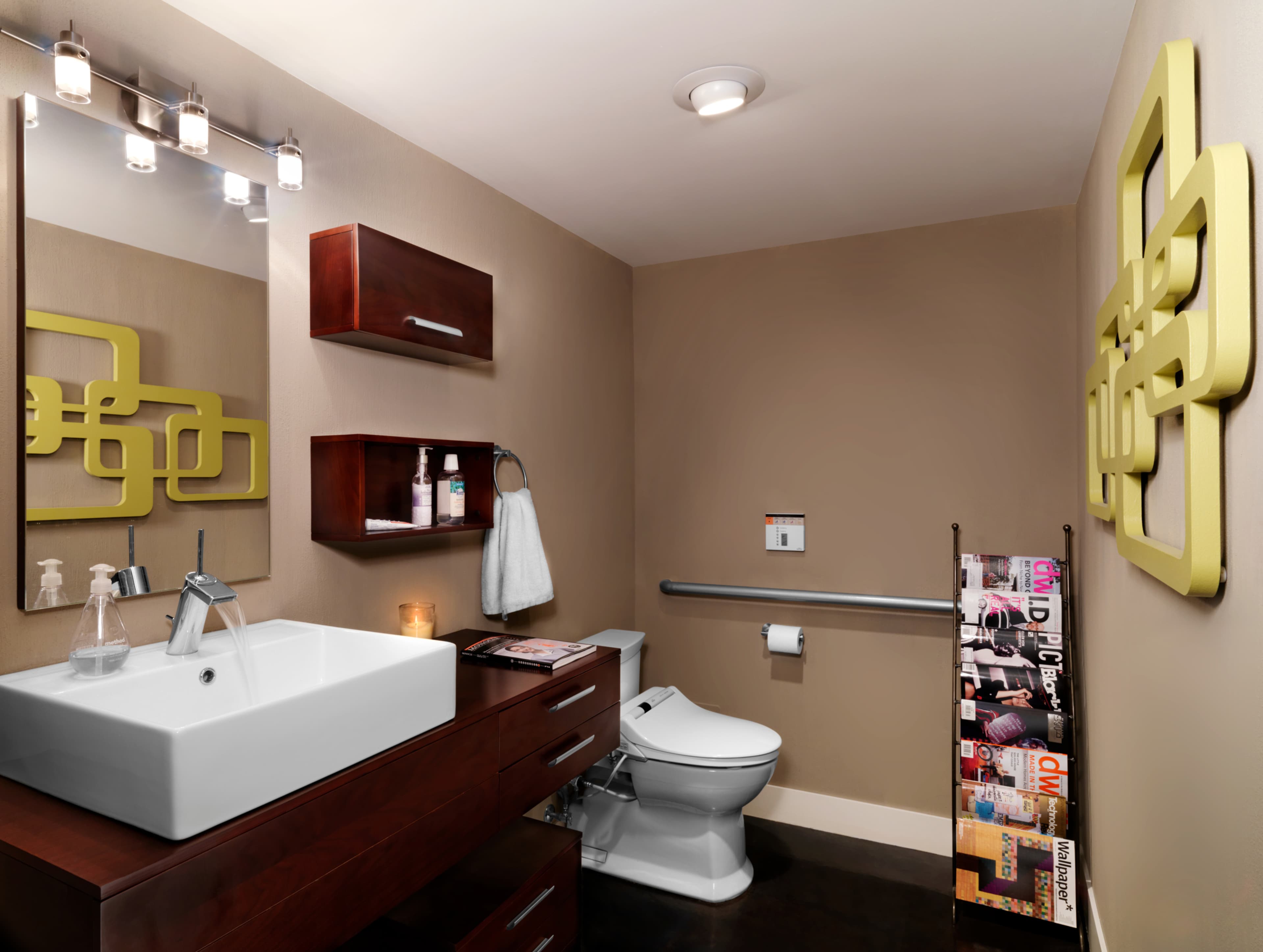 A modern bathroom with a white sink on wooden cabinetry, a toilet, and wall-mounted shelves, featuring a decorative wall piece and a magazine rack.