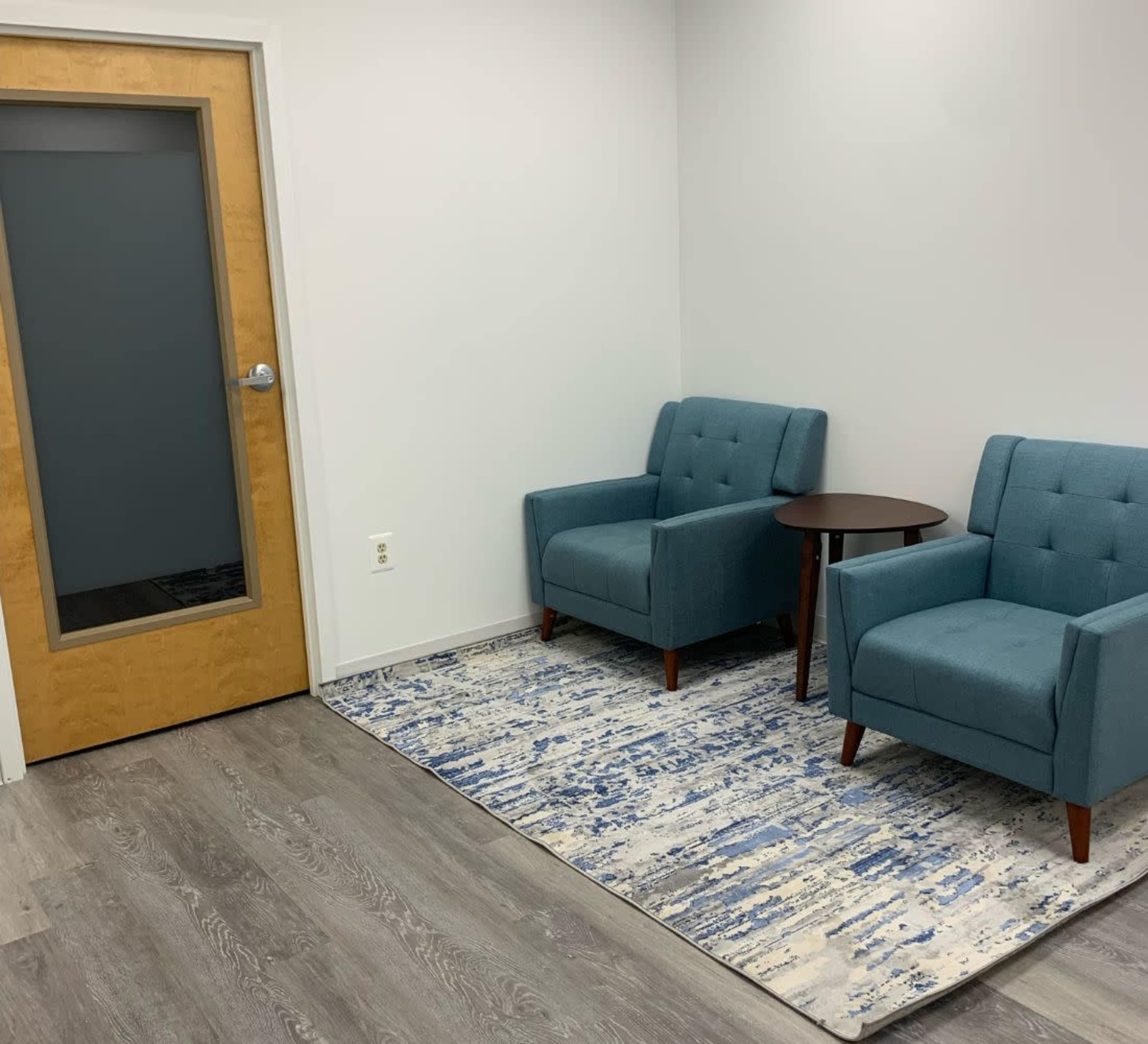 The image shows a small waiting area featuring two teal armchairs, a round table, and a patterned rug, with a wooden door leading to another room.
