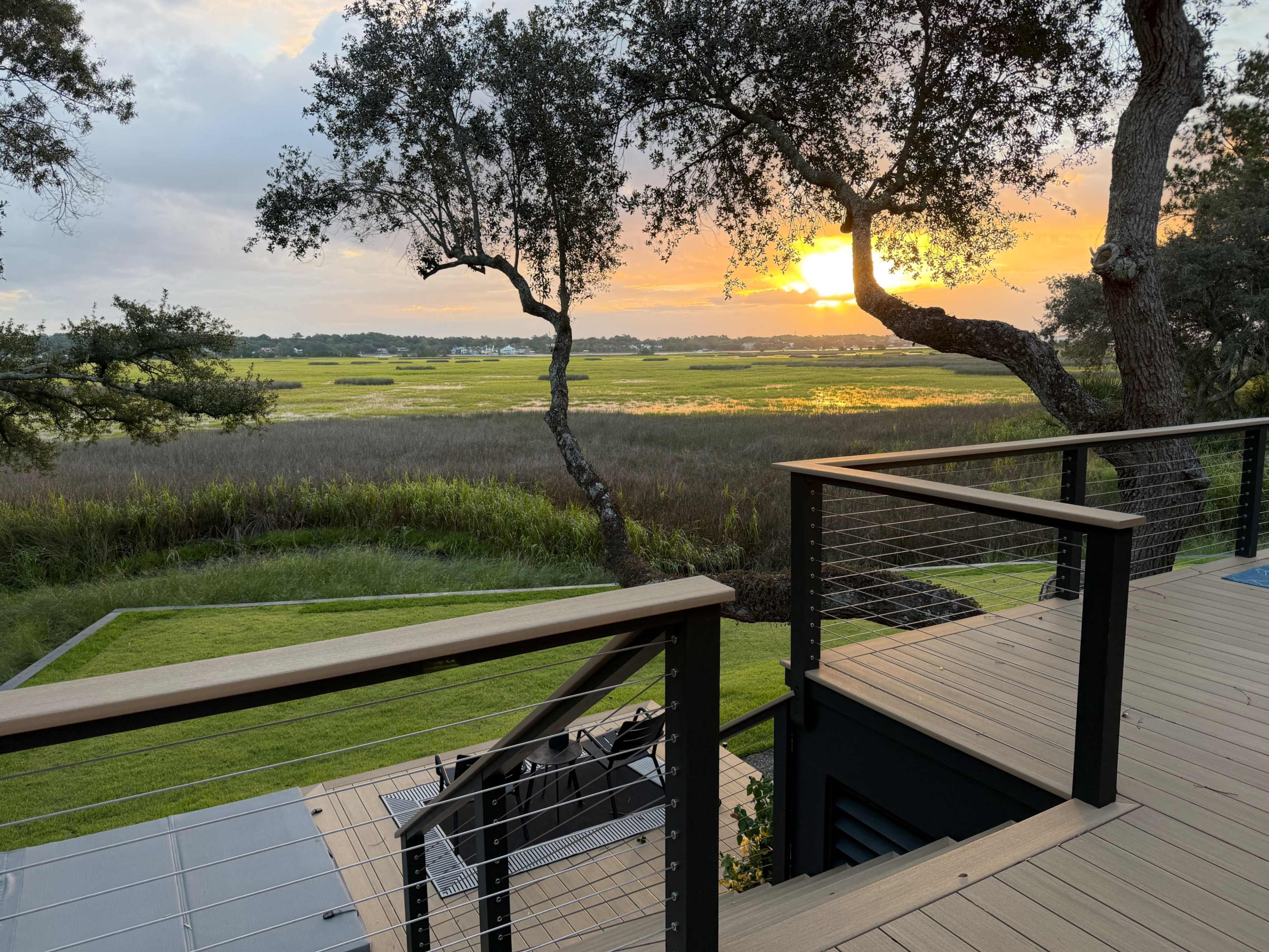 The image shows a wooden deck with metal railings overlooking a marshy landscape at sunset, with trees framing the view and the sun setting in the distance.