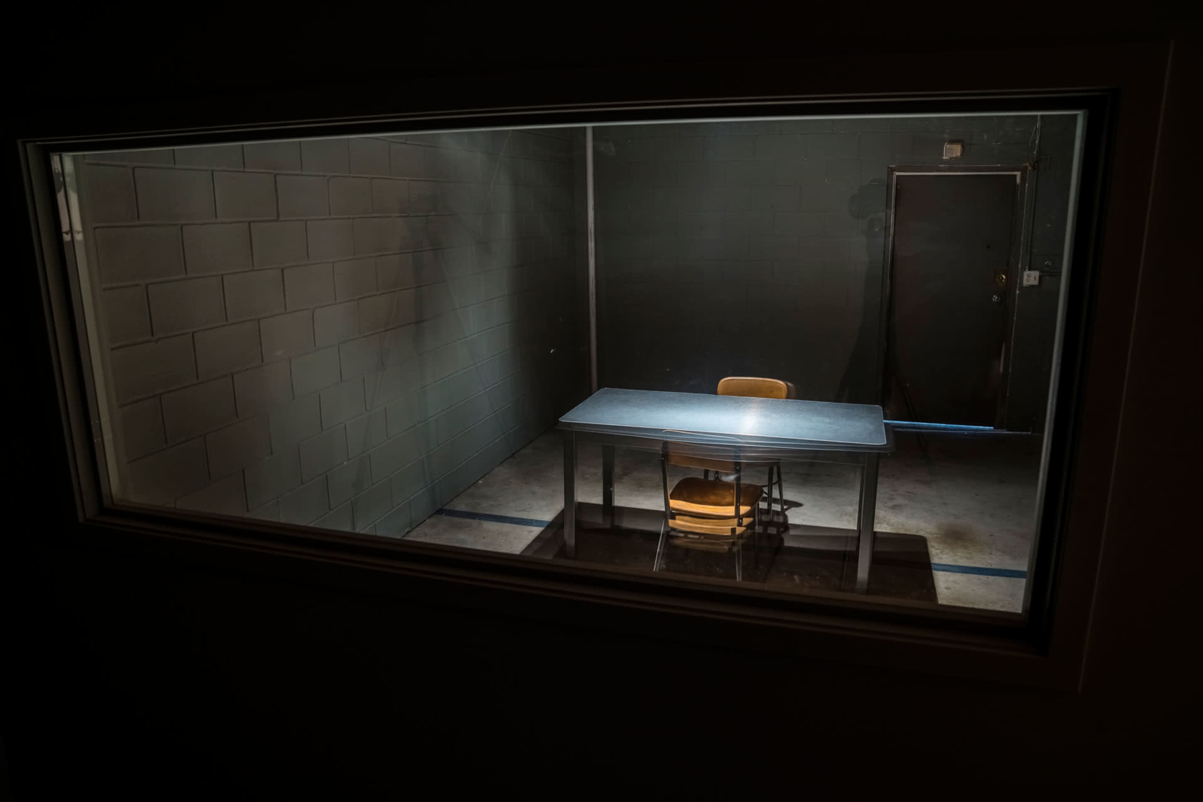 A vacant interrogation room is viewed through a window, featuring a single table and a chair against a bare wall.