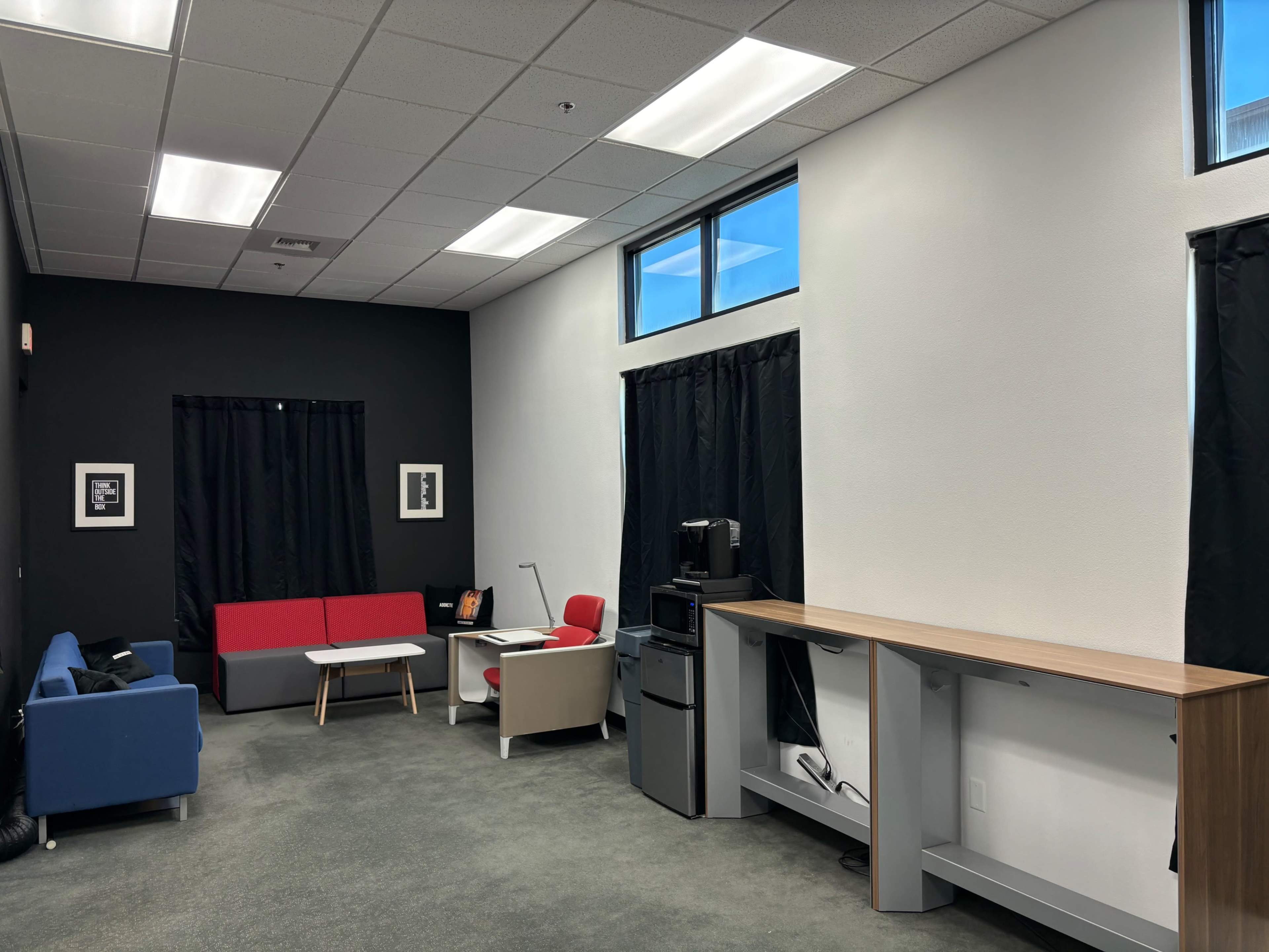 The image shows a modern office lounge featuring a blue couch, a red and grey seating area, a desk with a lamp, a printer, and a standing desk against a wall with black curtains.