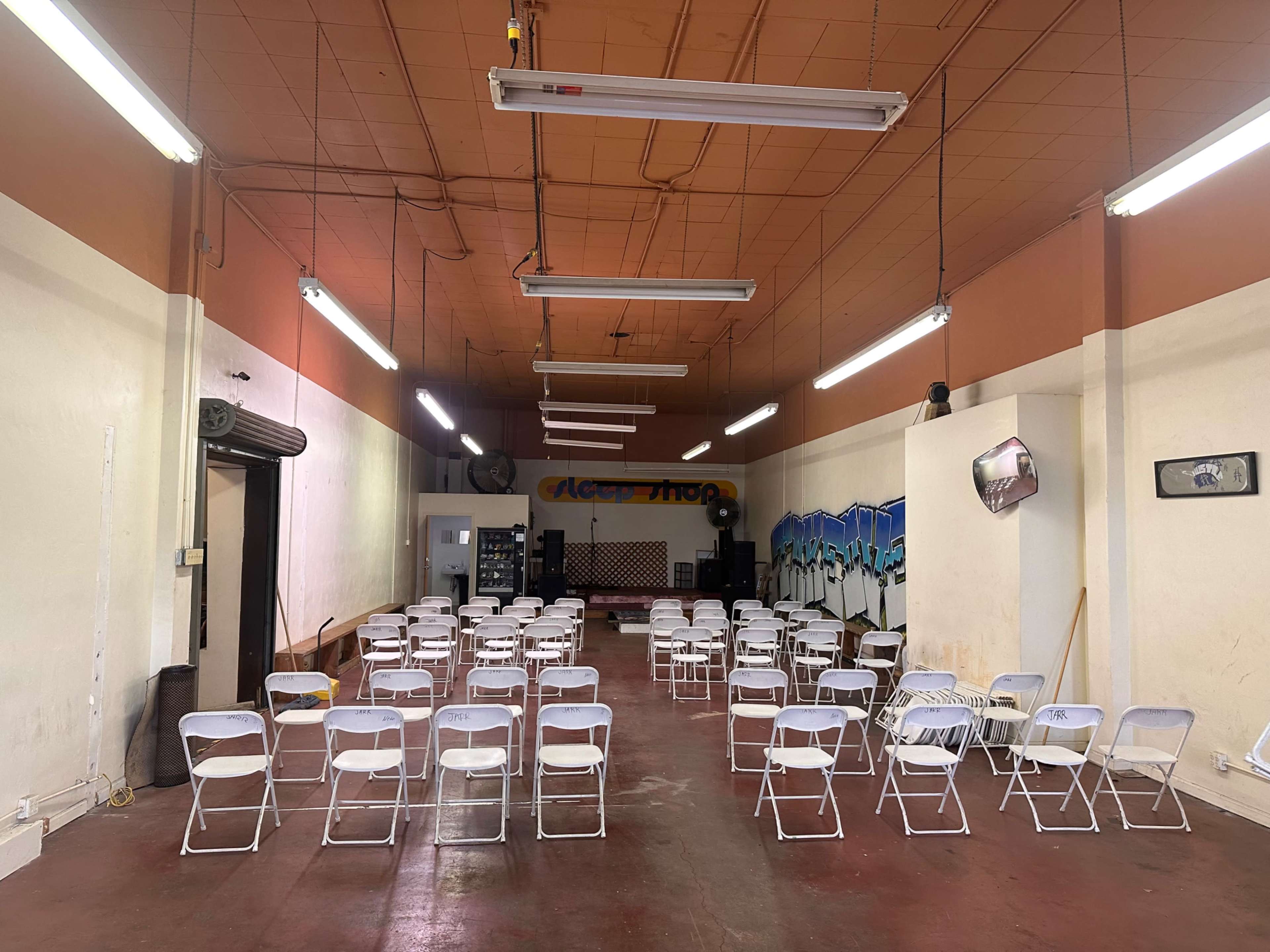 The image shows a spacious hall with rows of white folding chairs arranged facing a stage, illuminated by fluorescent lights.