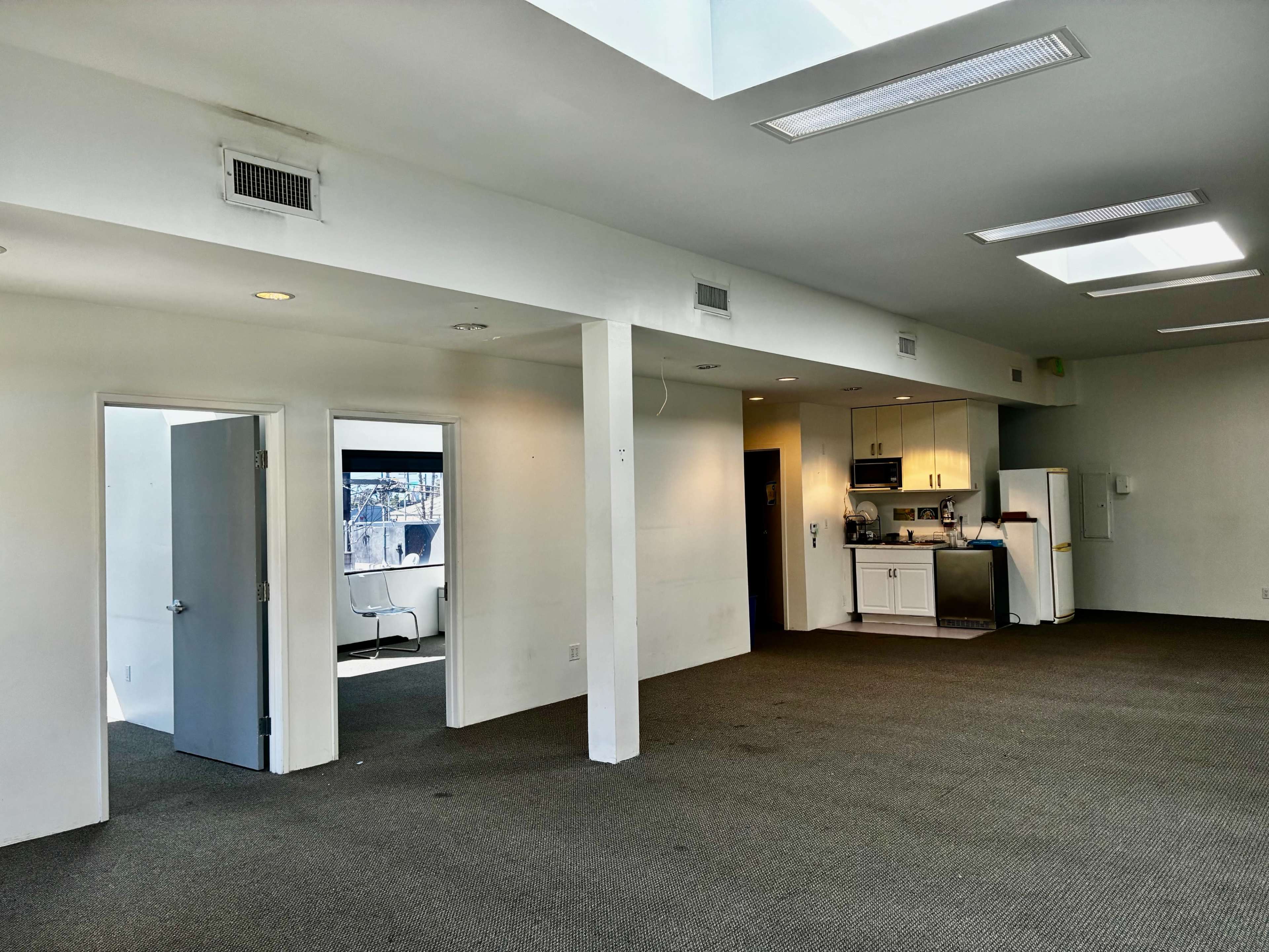 The image shows a spacious, empty room with a kitchen area, two doors, and skylights allowing natural light.