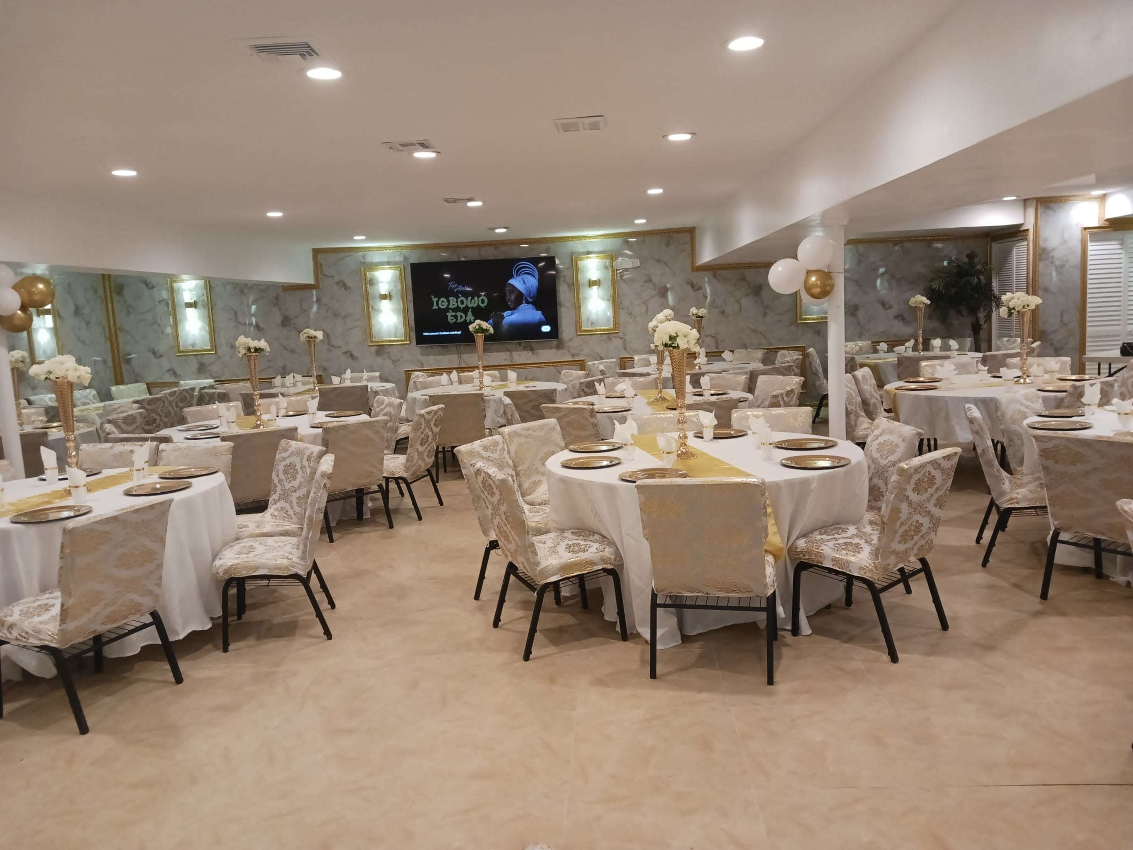 A banquet hall with round tables dressed in white tablecloths and gold accents, surrounded by upholstered chairs, and featuring wall decorations and a large screen at the front.