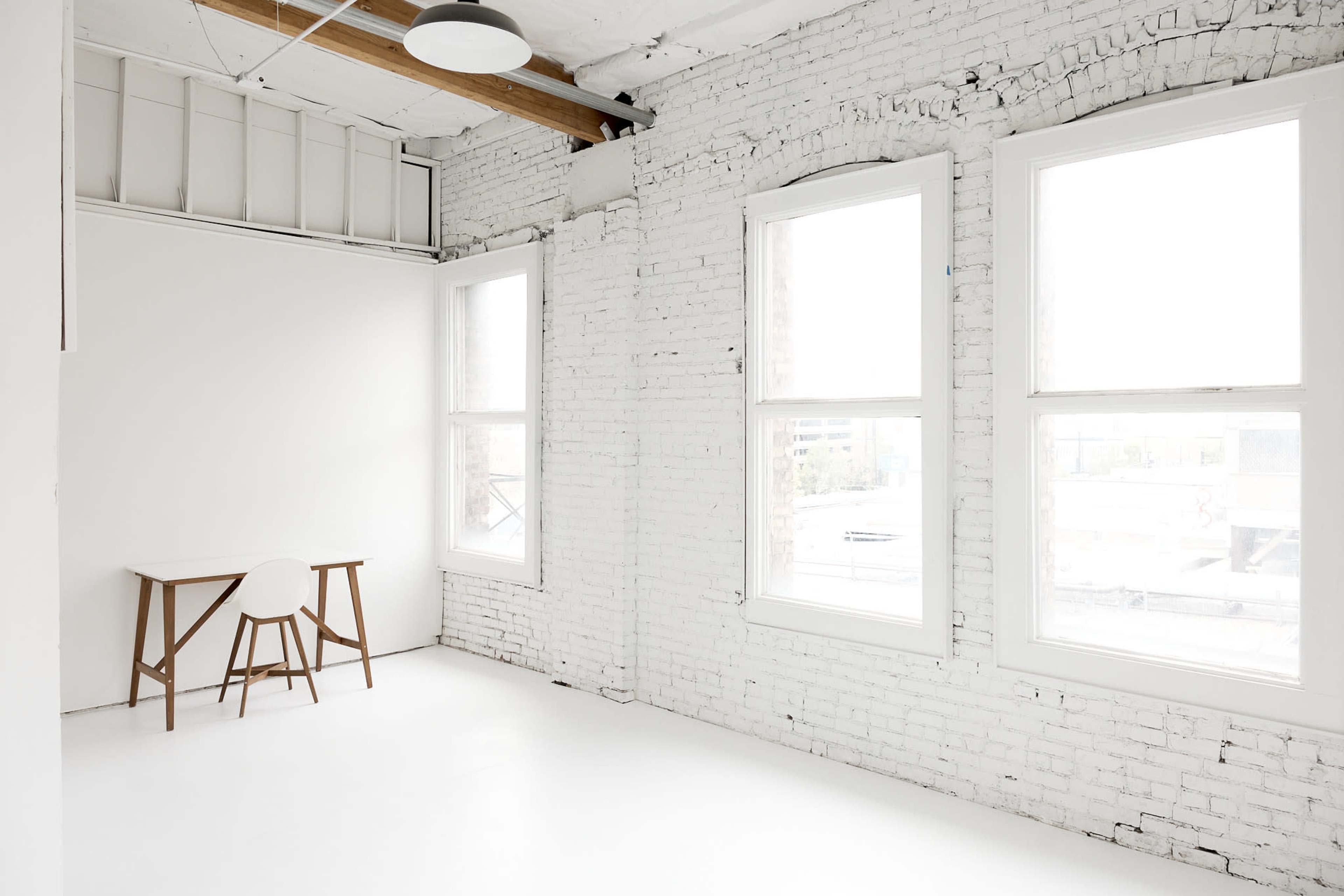 The image shows a bright, minimalistic room with white brick walls, a simple wooden table, and a round chair near large windows.