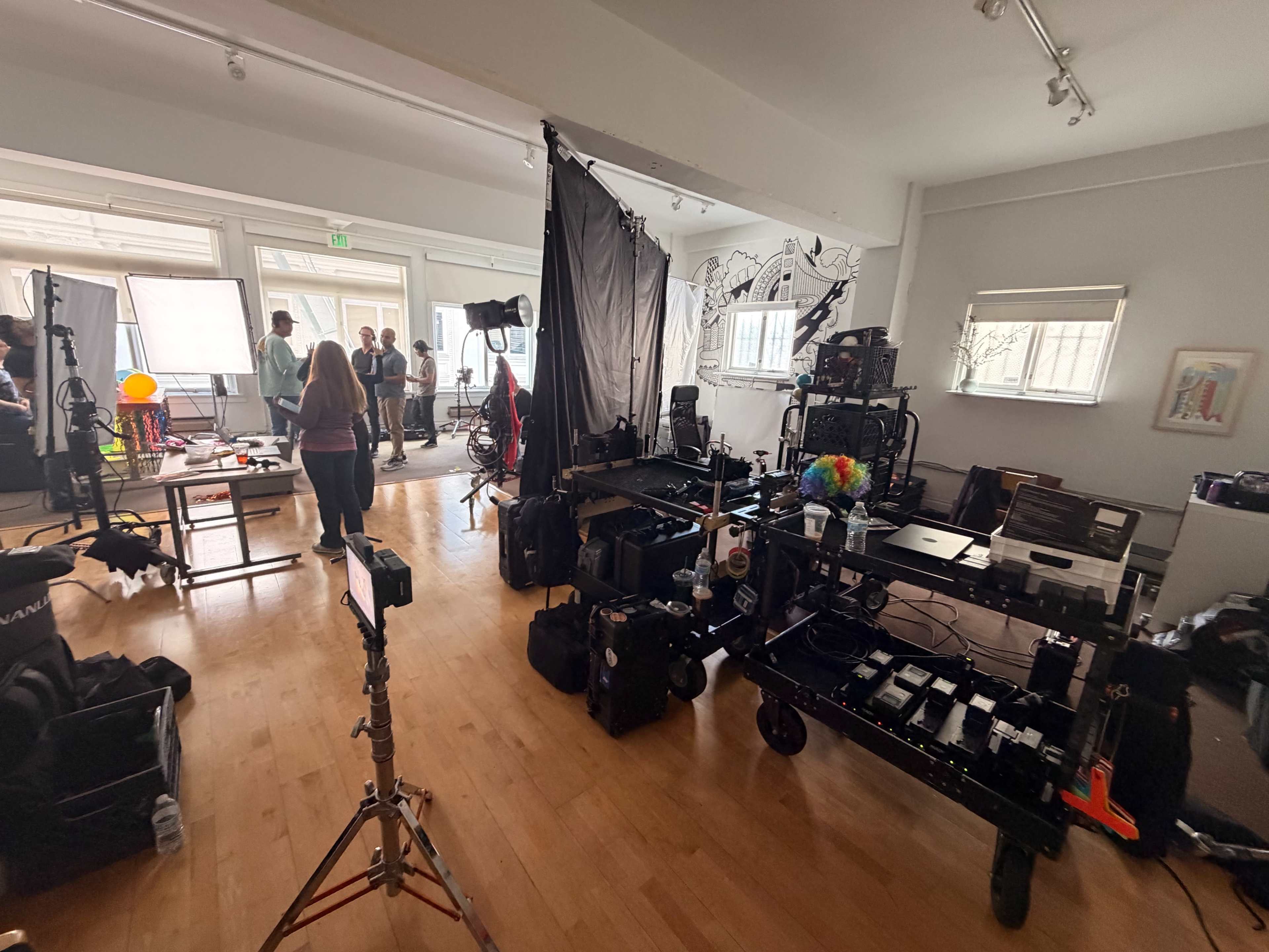 The image shows a photography studio filled with various lighting equipment, a camera setup, and several people engaged in conversation.