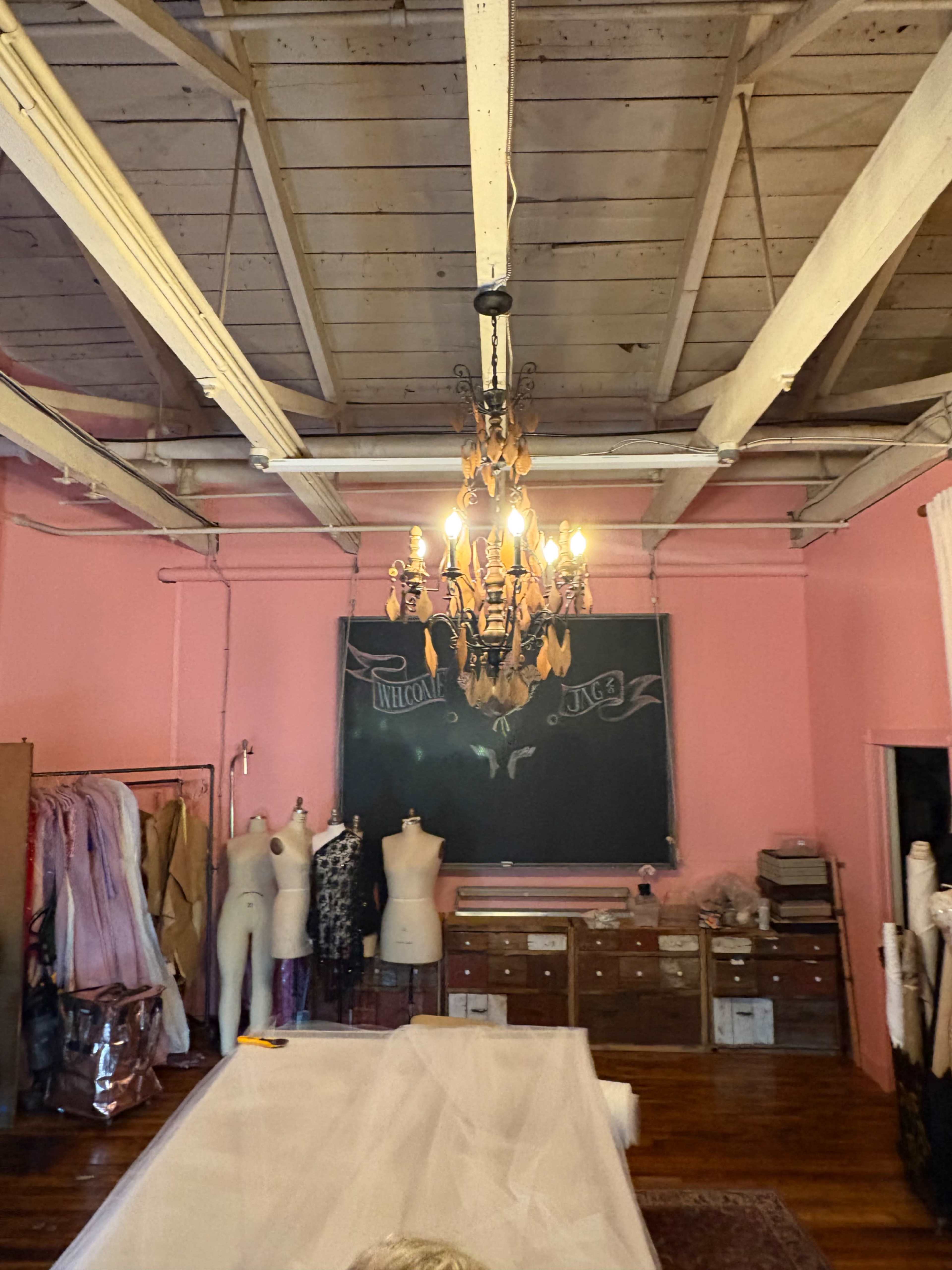 The image shows a brightly lit fabric studio featuring a chandelier, a pink backdrop, sewing mannequins, and organized rolls of fabric.